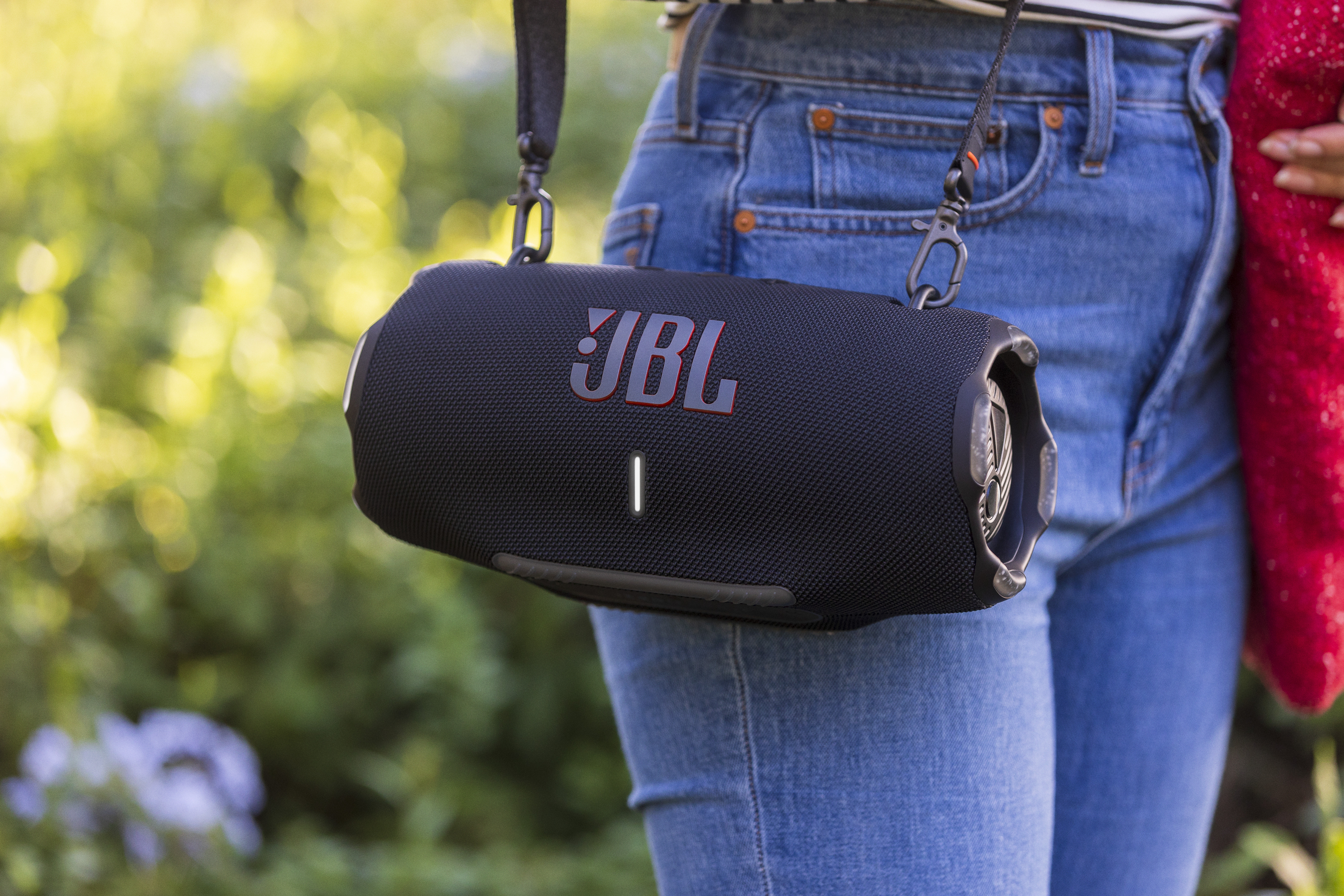 Una persona con jeans e un altoparlante JBL nero. L'altoparlante è nero con scritte rosse e una tracolla.