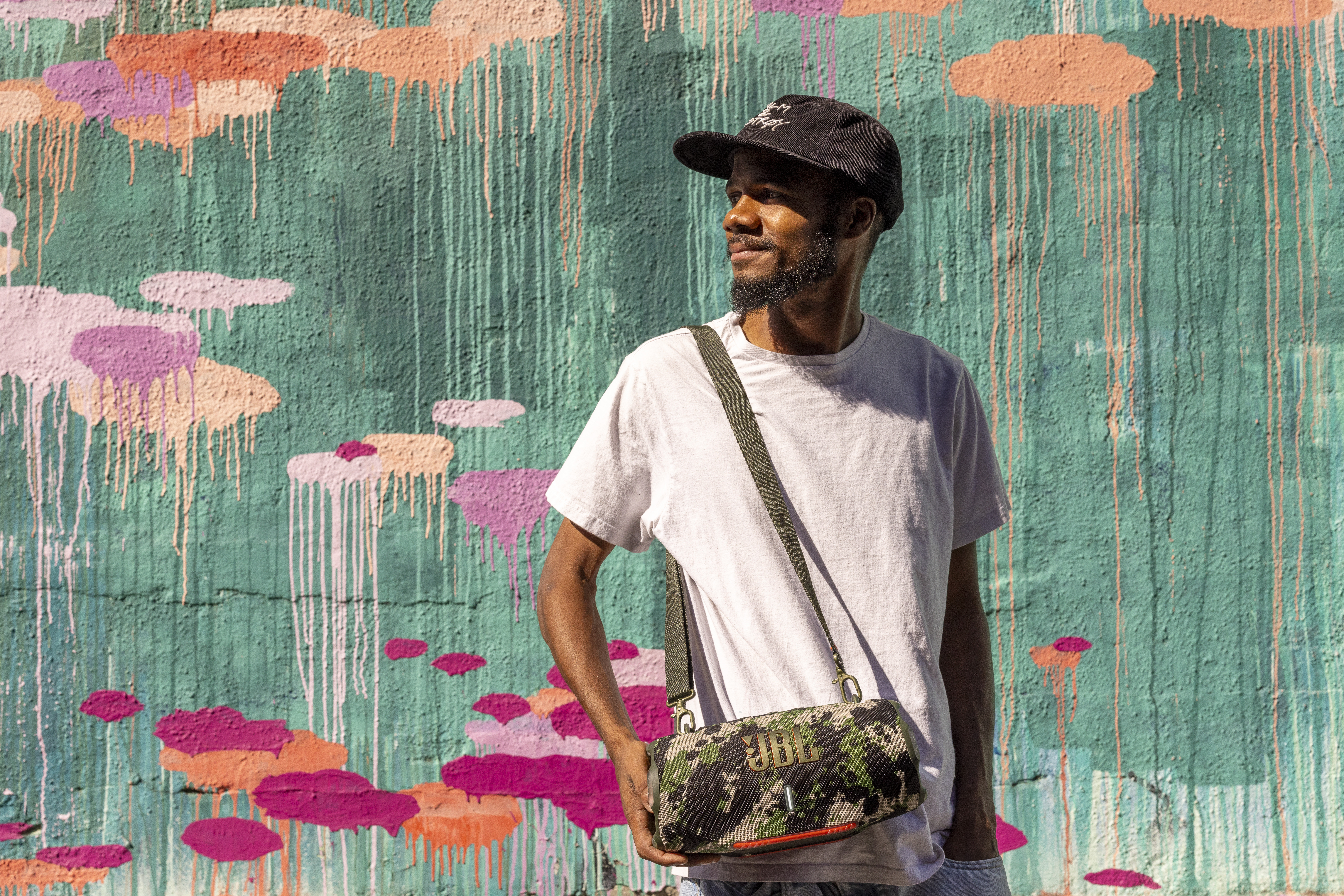 Un homme avec une enceinte JBL camouflage sur l'épaule, devant un mur de graffitis colorés.