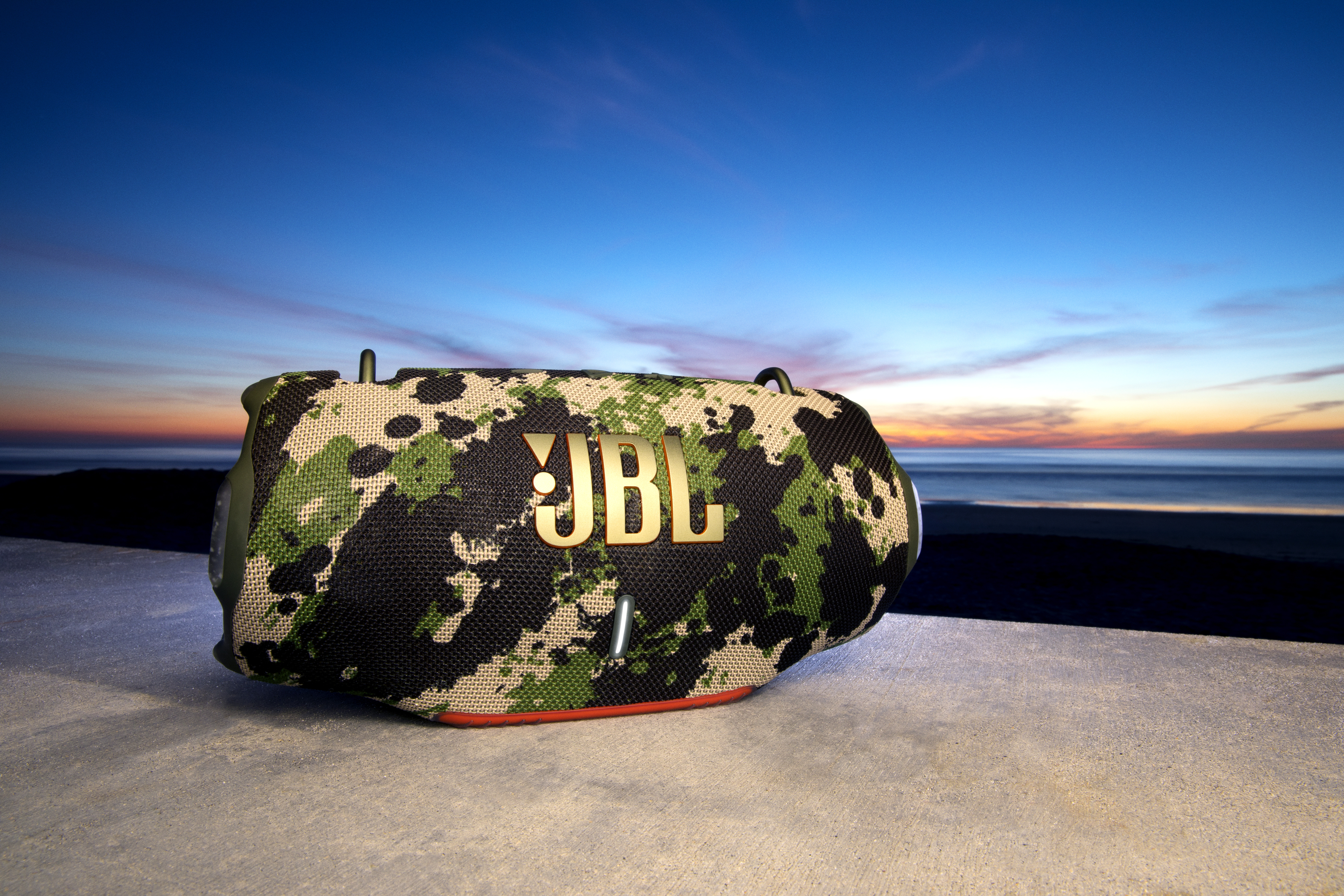 Une enceinte JBL sur une plage au coucher du soleil. Motif camouflage, accents orange.