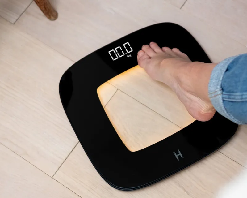 Fekete mérleg, láb a mérlegen, 00,0 kg-ot mutat. Világos fa felület.