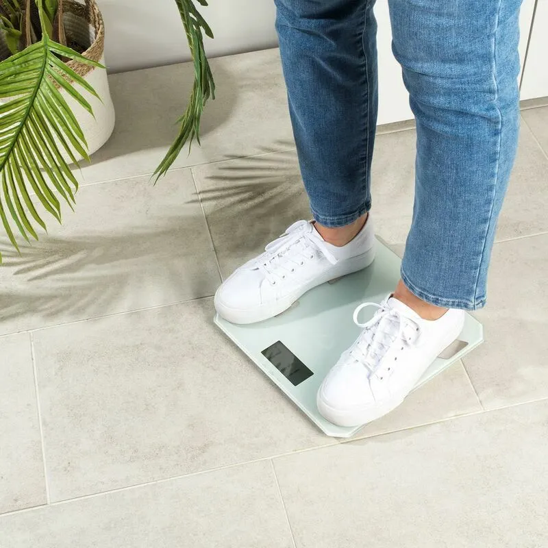 Személy fehér testmérlegen áll, 0,0 kg-ot mutat. Fehér cipő, farmer, növények a háttérben. Salter márka.