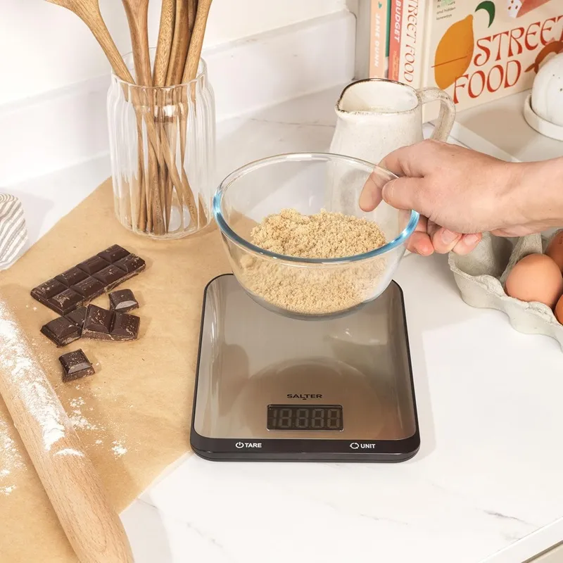 Egy kéz barna cukorral teli tálat tart egy mérlegen. Csokoládé, tojás és konyhai eszközök vannak az asztalon.