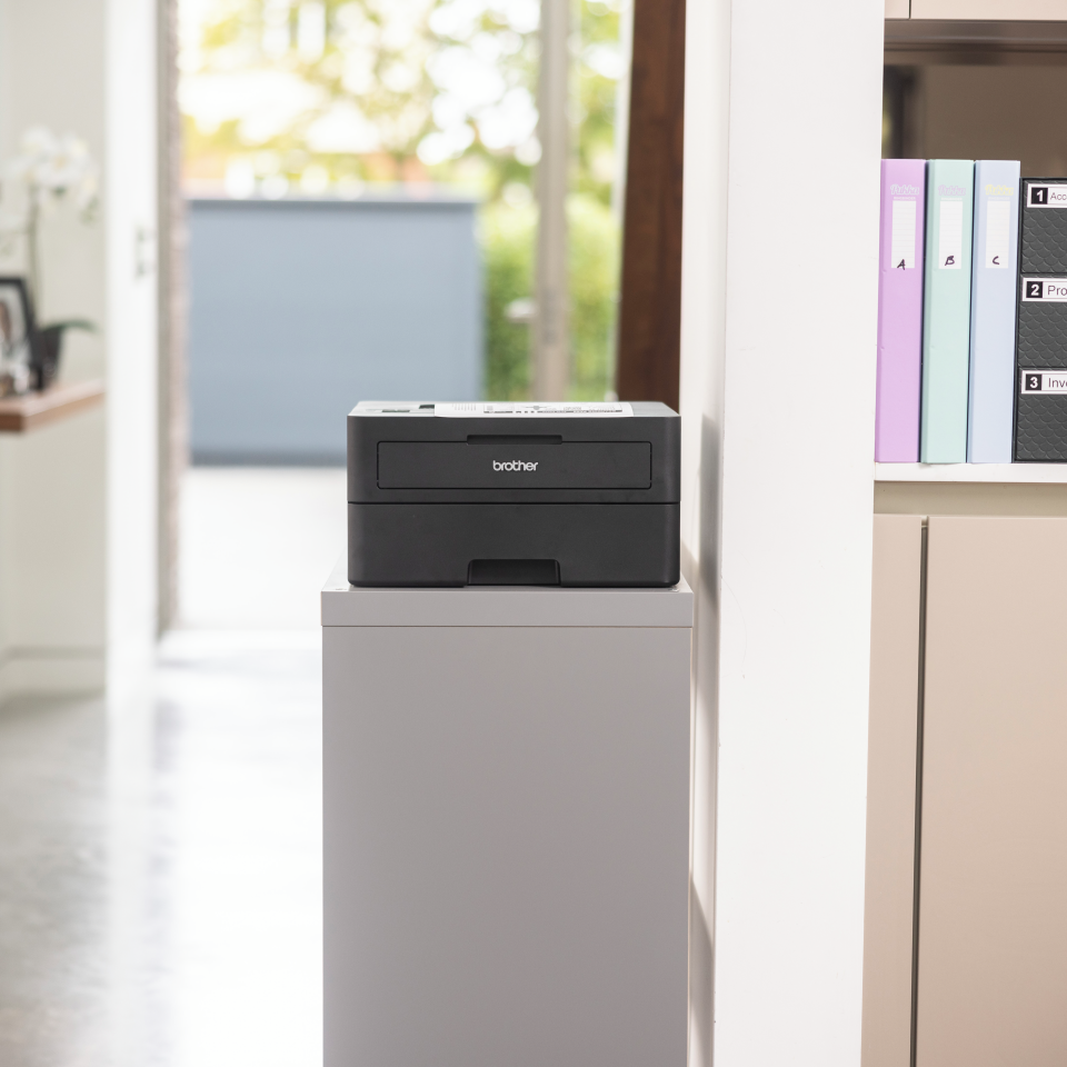 Czarna drukarka na szarej szafce. Białe ściany, drzwi i półka z segregatorami są widoczne.