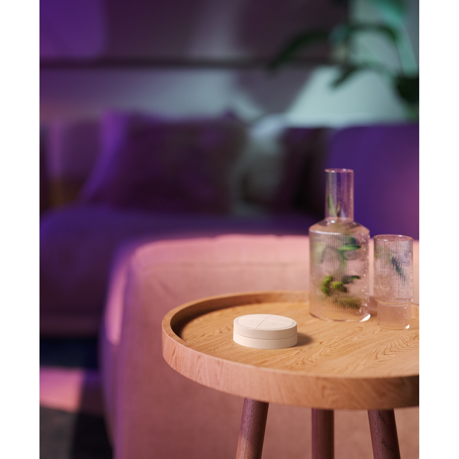 Un interruttore Philips Hue rotondo è appoggiato su un tavolo di legno accanto a un bicchiere e un vaso.