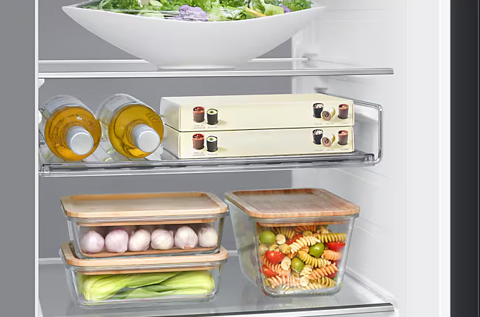 Wnętrze lodówki z półkami. Zawiera butelki, pudełka, szklane pojemniki z makaronem, czosnkiem i sałatą. Sałatka w białej misce.