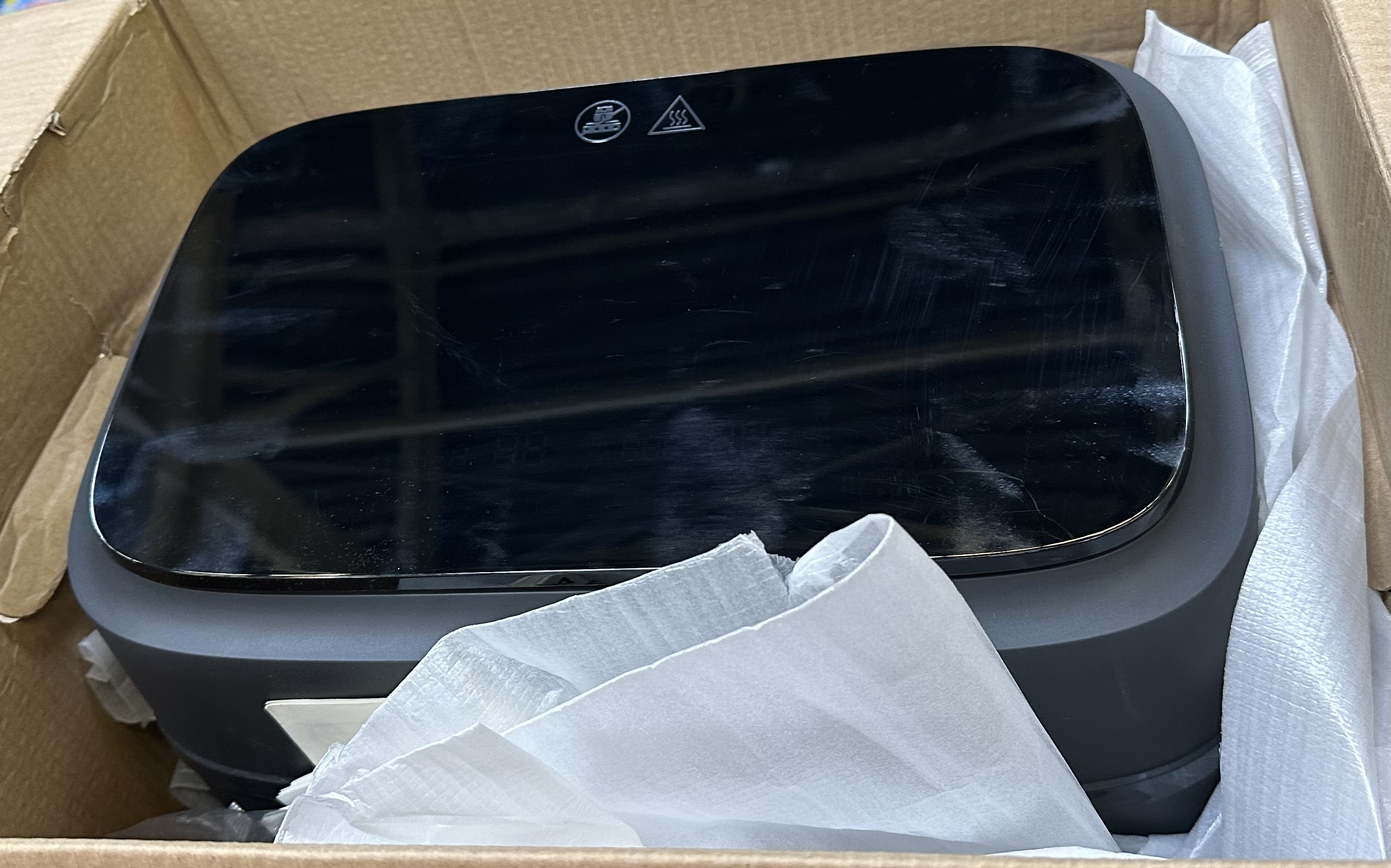 Czarna frytkownica spoczywająca wewnątrz kartonowego pudełka, z błyszczącą czarną pokrywą.