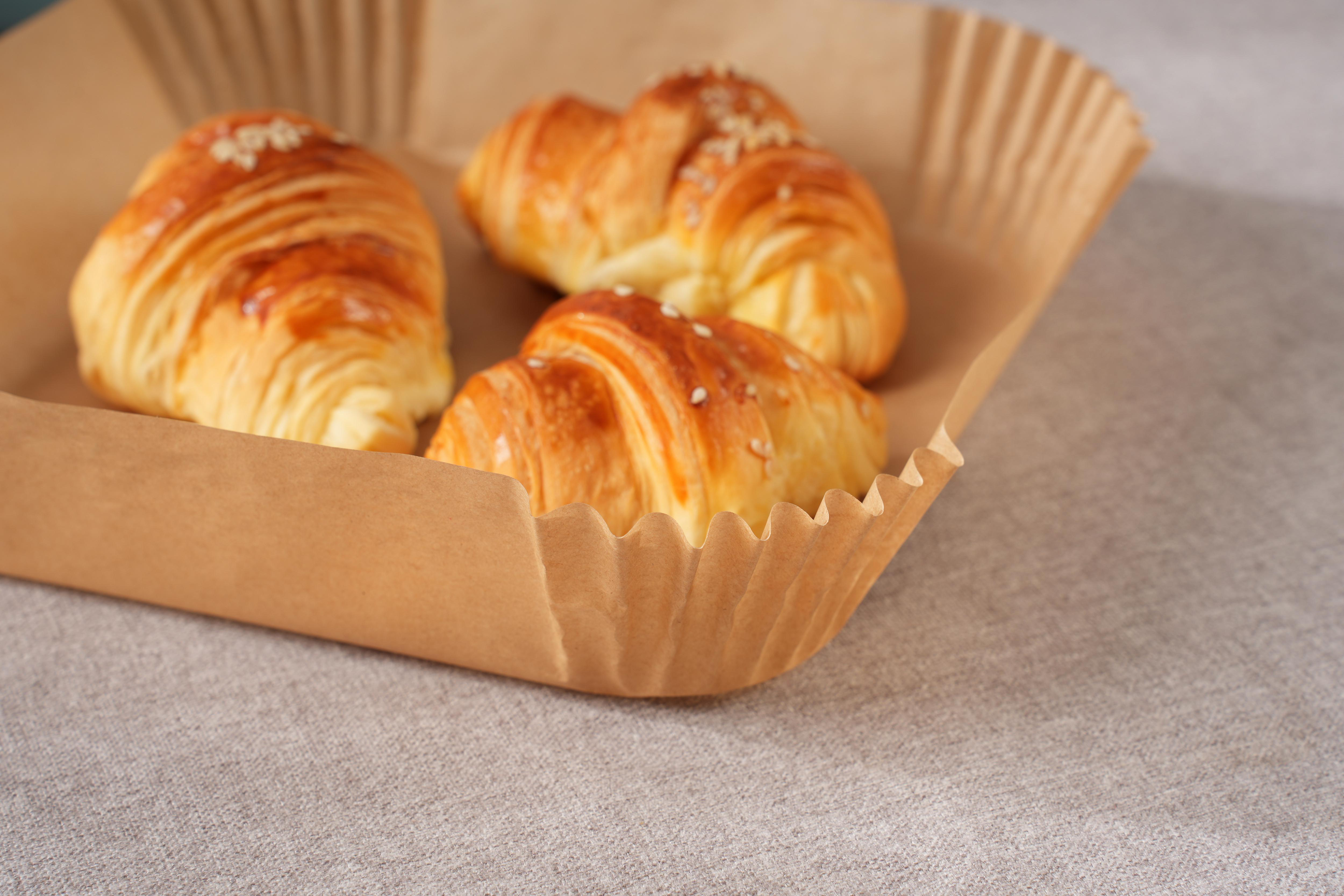 Drei goldbraune Croissants in einer braunen Papierbackform auf einem grauen Tisch.