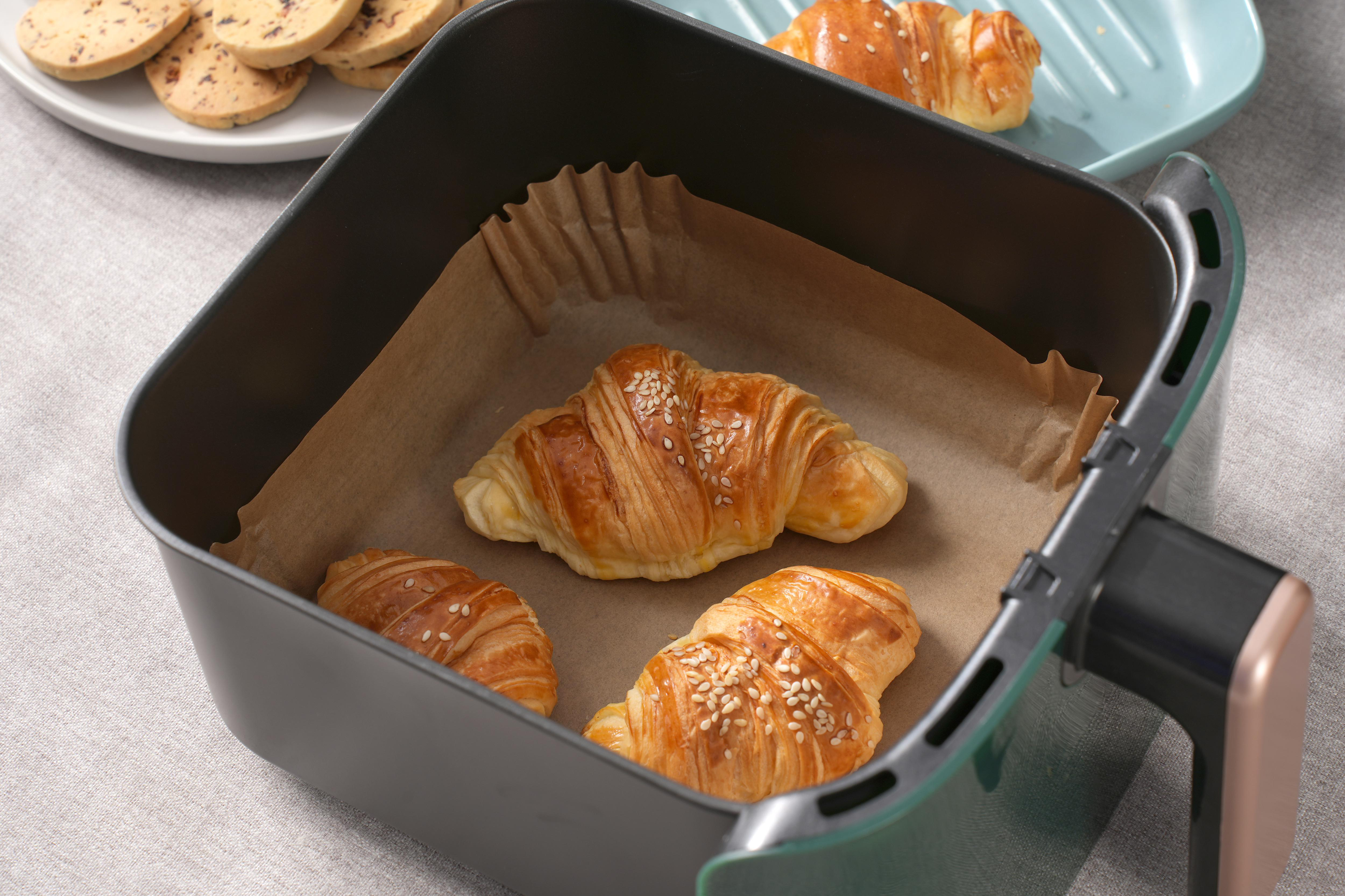 Drei goldbraune Croissants mit Sesam in einer Airfryer. Im Hintergrund Gebäck und eine blaue Platte.