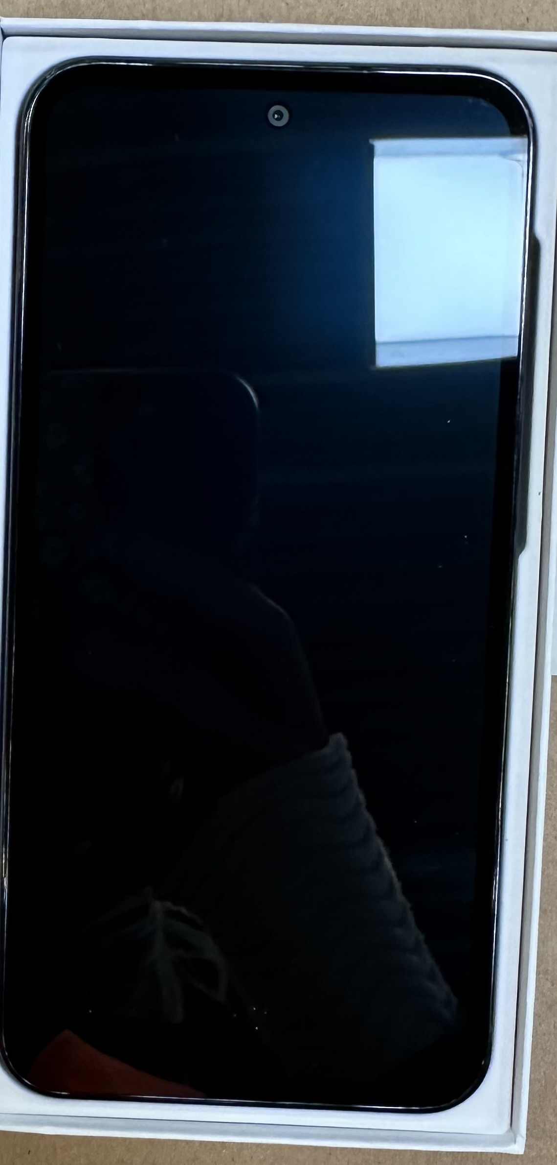 Czarny smartfon w białym pudełku, aparat na środku u góry, ekran odbija światło.