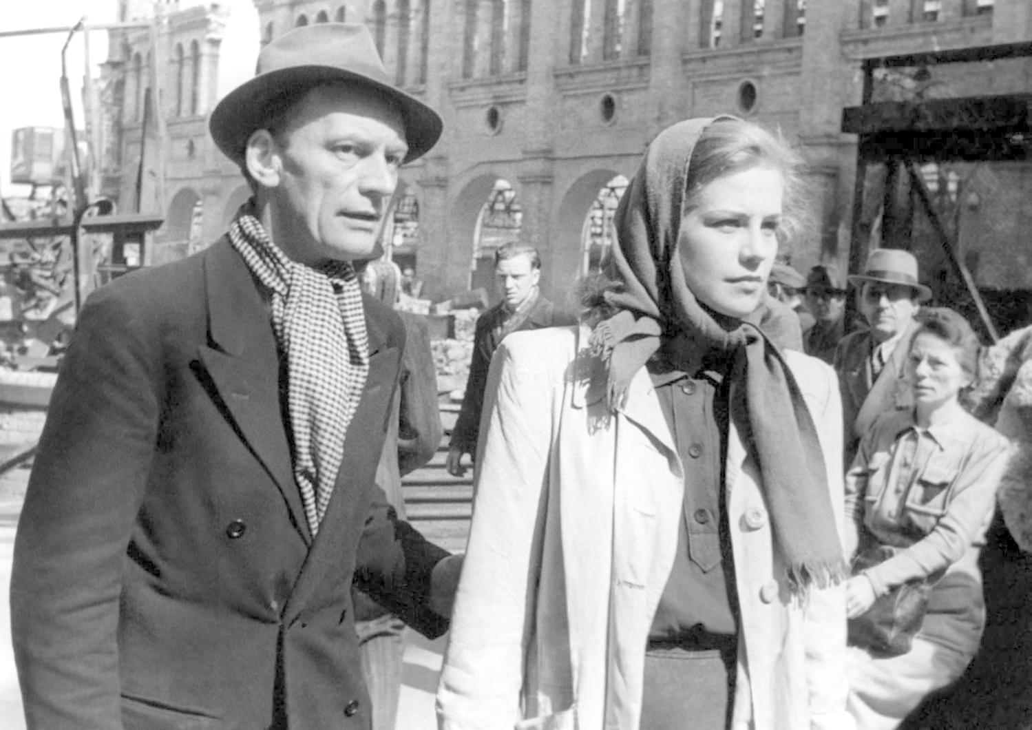 Ein Mann und eine Frau gehen an einem zerstörten Gebäude vorbei. Der Mann trägt Hut und Schal, die Frau Kopftuch.