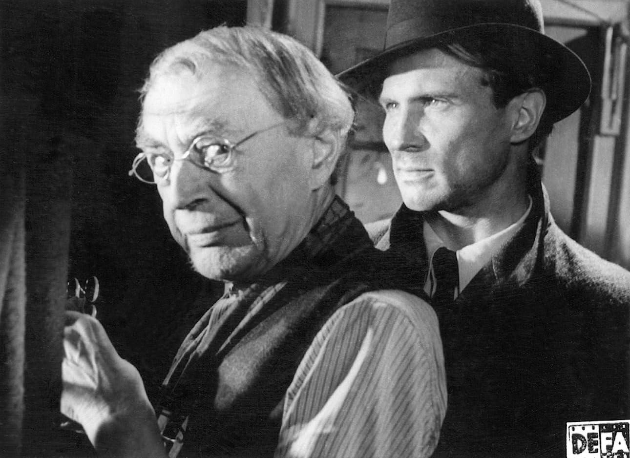 Ein älterer Mann mit Brille und ein junger Mann mit Hut schauen in die gleiche Richtung. Beide sind in einem Raum.