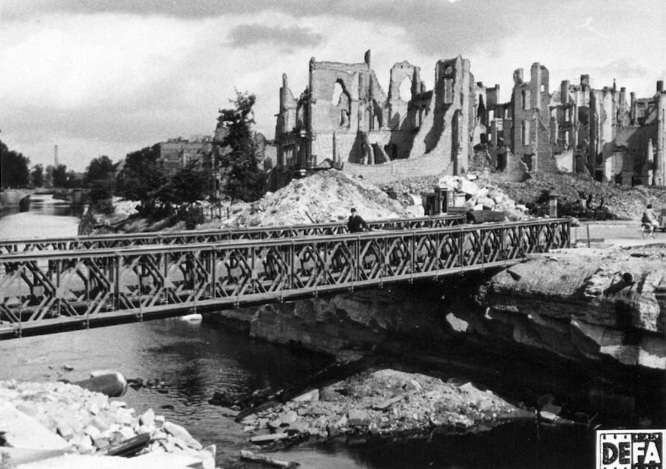 Brücke über einem Fluss mit zerstörten Gebäuden im Hintergrund.