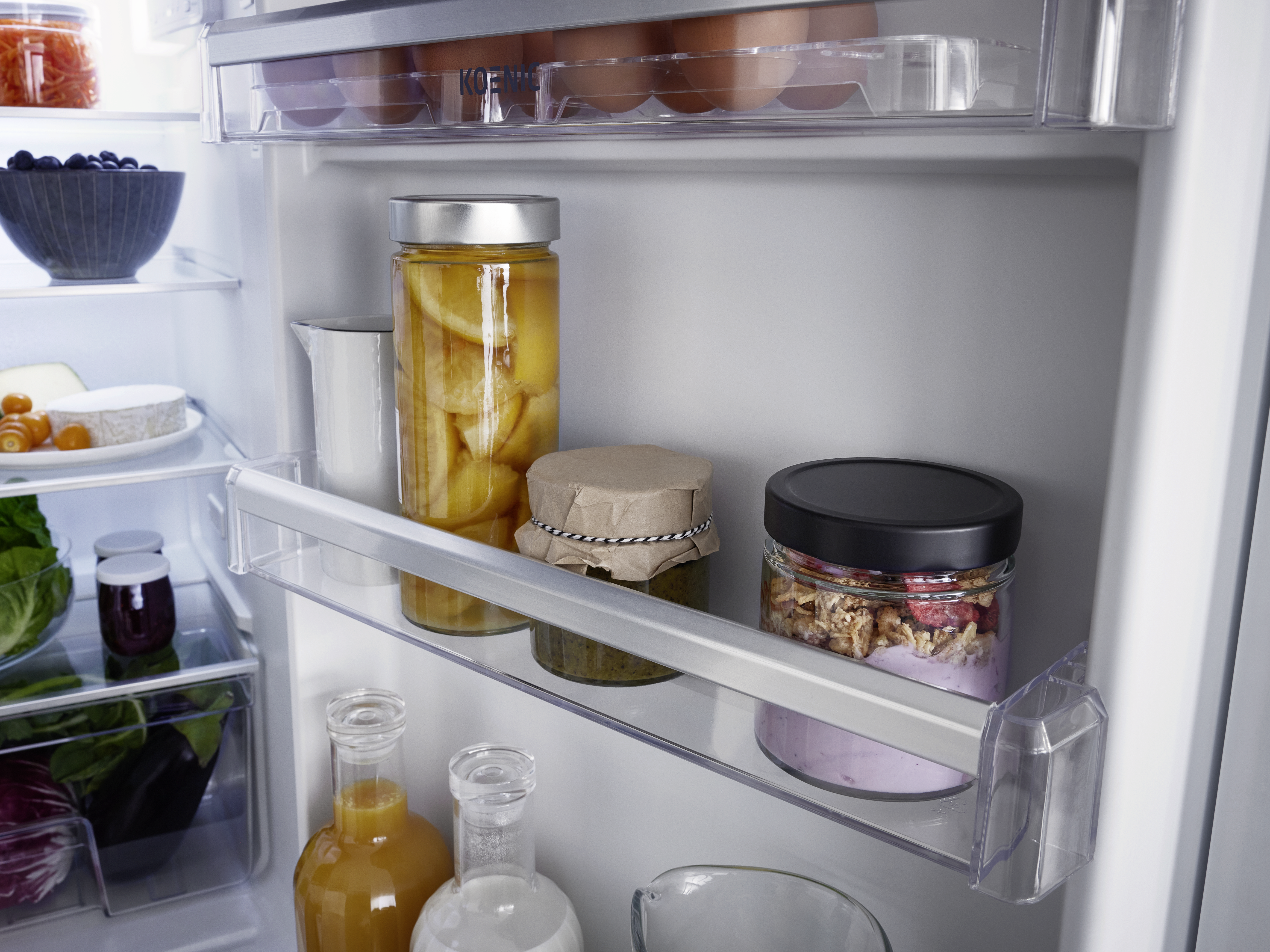 Der Kühlschrank ist geöffnet, mit Lebensmitteln gefüllt. Auf den Regalen sind Gläser, Eier, Obst und Gemüse zu sehen.