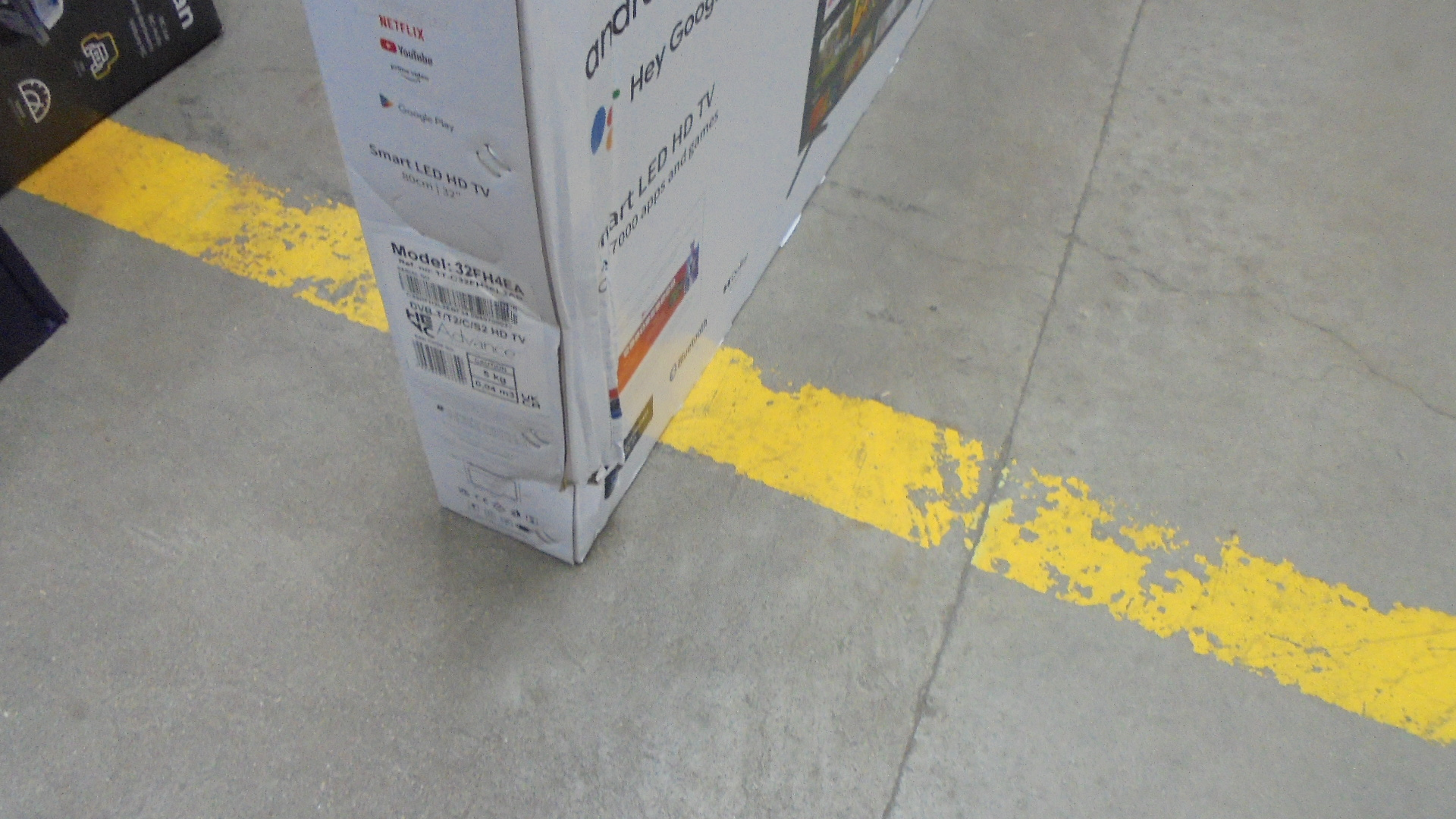 Białe pudełko z nadrukami, stojące na szarej podłodze, z żółtą linią.