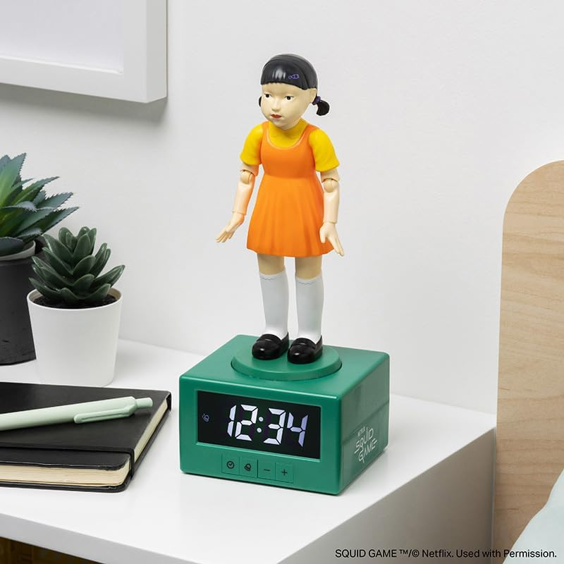 Squid Game figura ébresztőóra egy éjjeliszekrényen. Az idő 12:34. Növények és fal a háttérben.