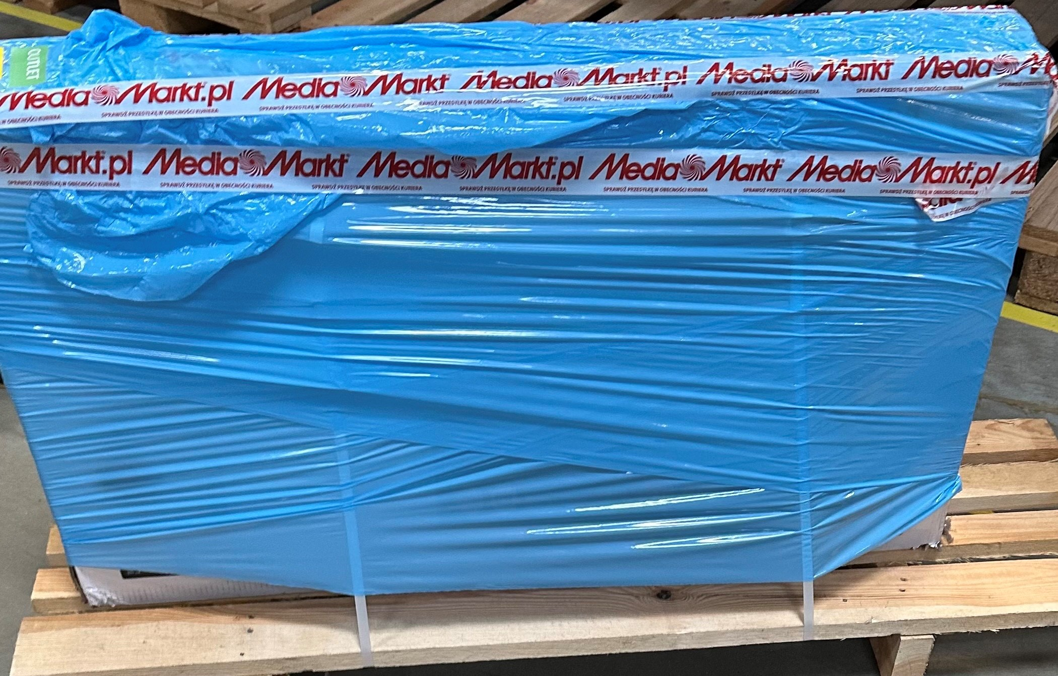 Niebieska folia na drewnianej palecie. Widoczna taśma Media Markt.pl.