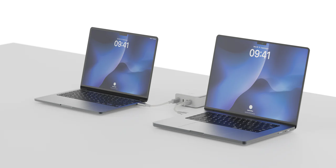 Dos portátiles conectados a un concentrador USB-C en una superficie blanca, ambos mostrando una pantalla de bloqueo.