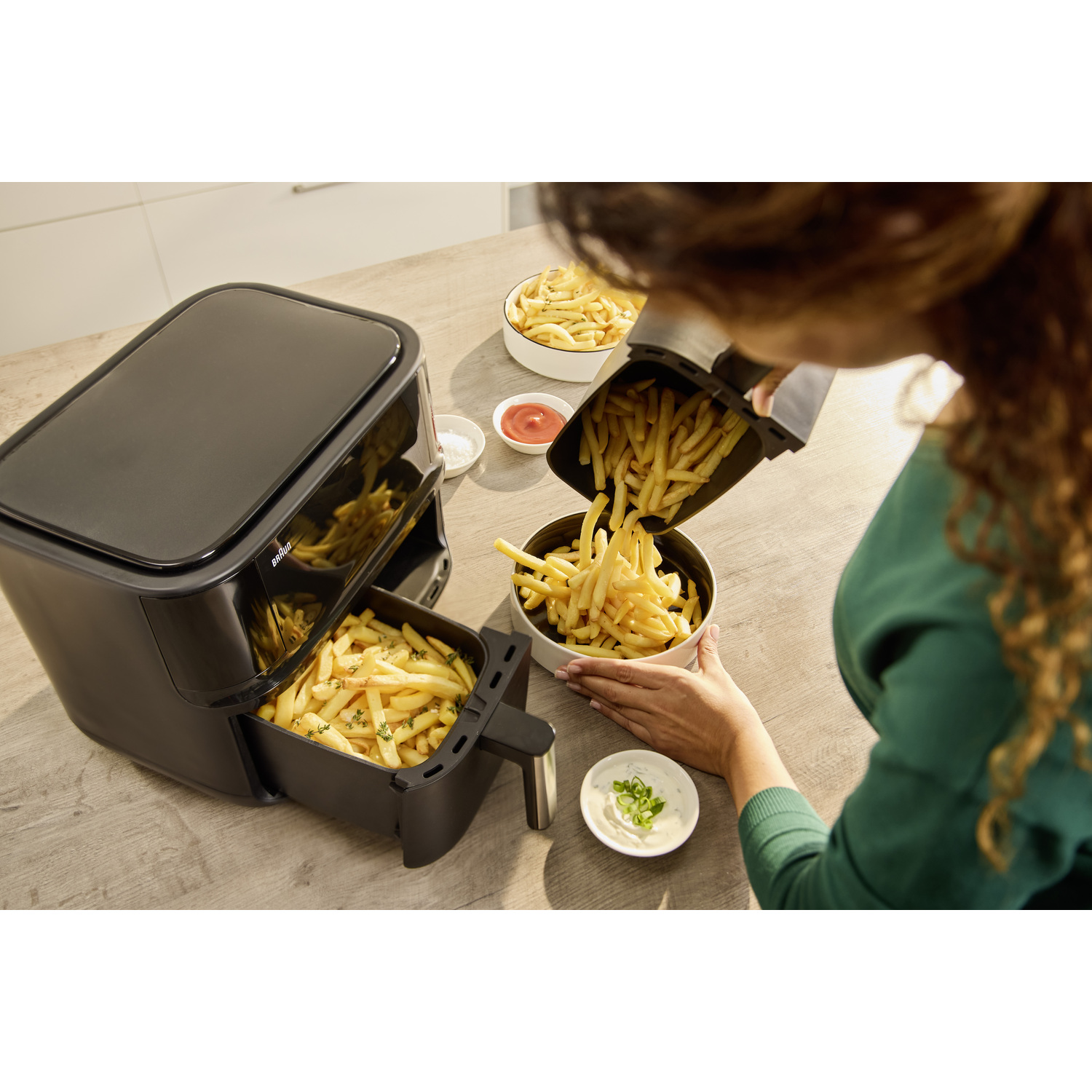 Een persoon giet frietjes uit een airfryer in een kom. Airfryer en kommen met frietjes staan op een houten tafel met kruiden.