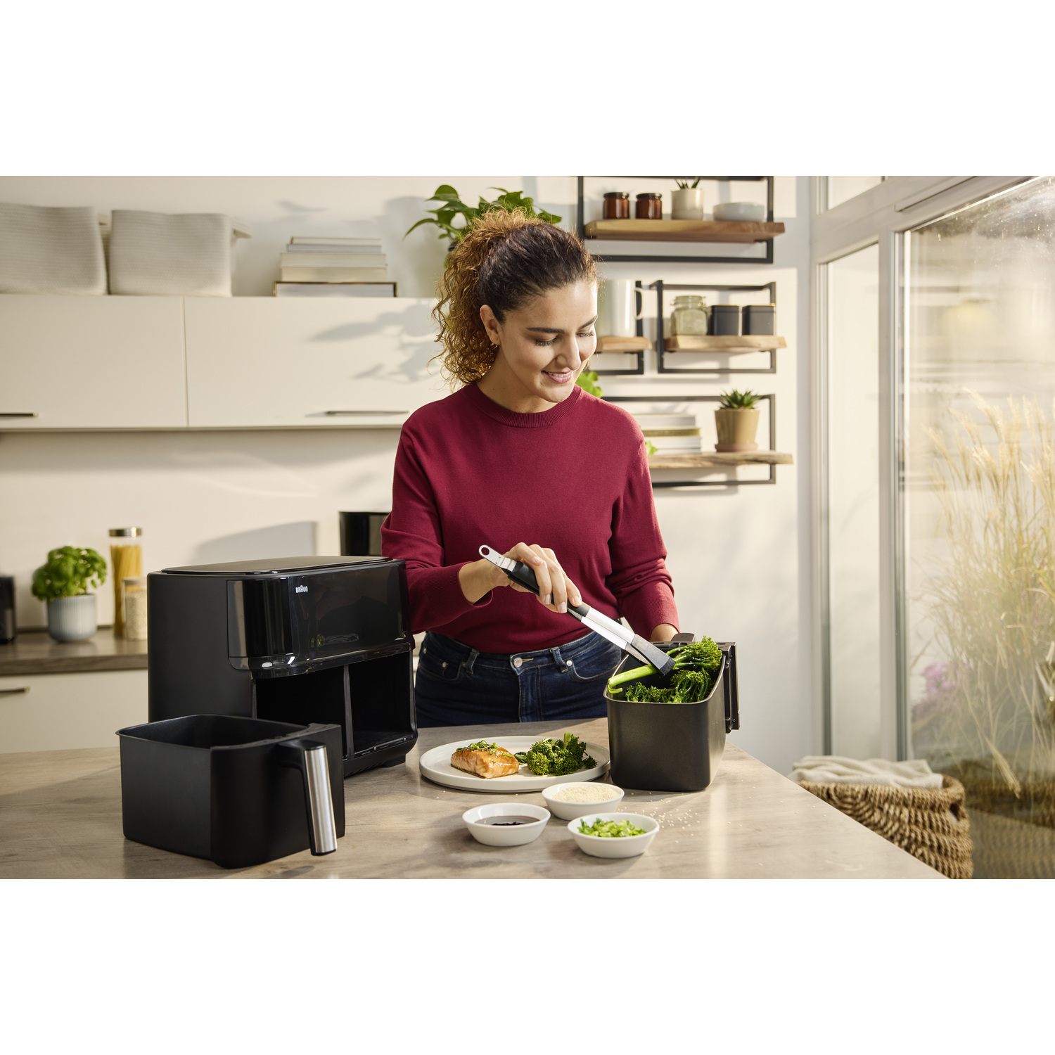 Vrouw in keuken met tang voor groenten, airfryer, eten op bord en kommen op tafel.