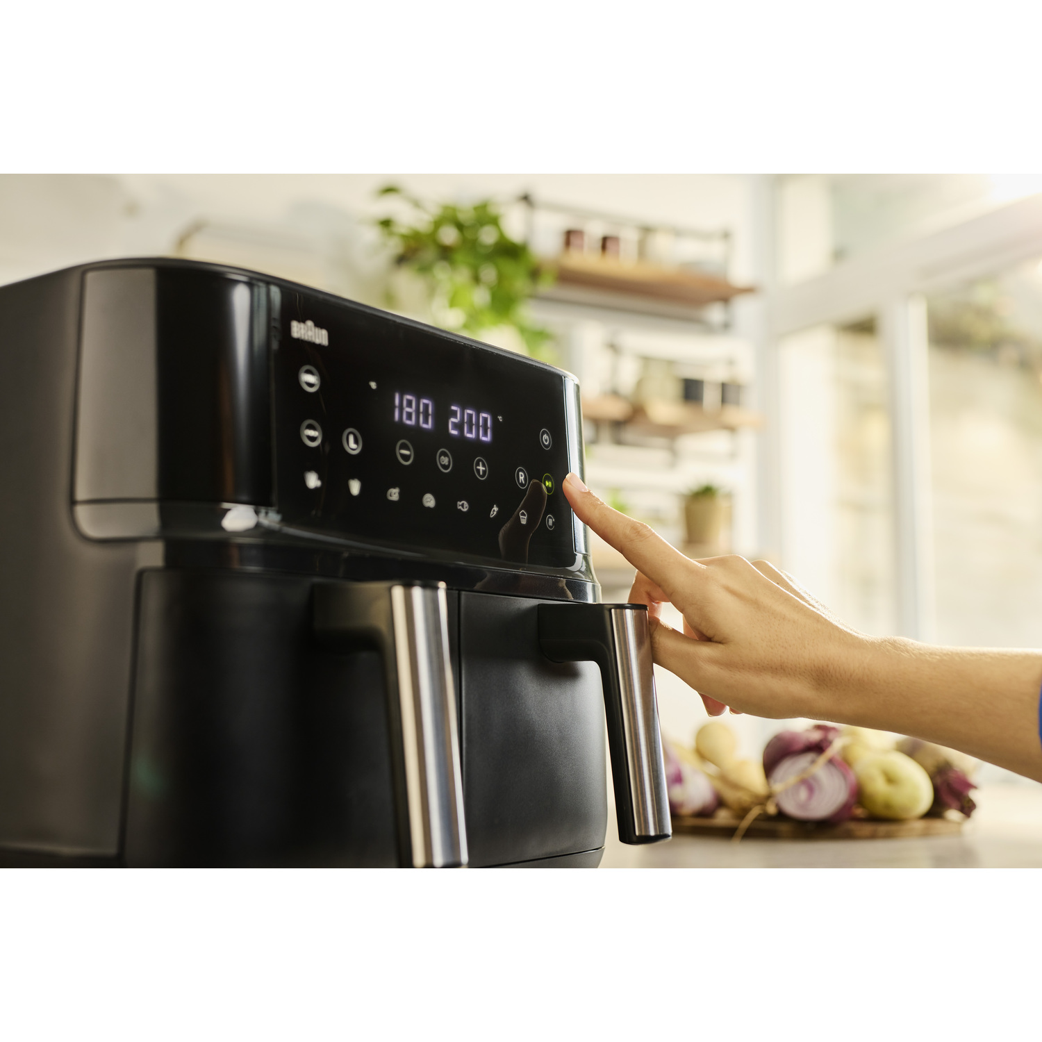 Een hand drukt op een knop van een zwarte airfryer. Het display toont 180 en 200. Groenten zoals uien en aardappelen zijn op de achtergrond.