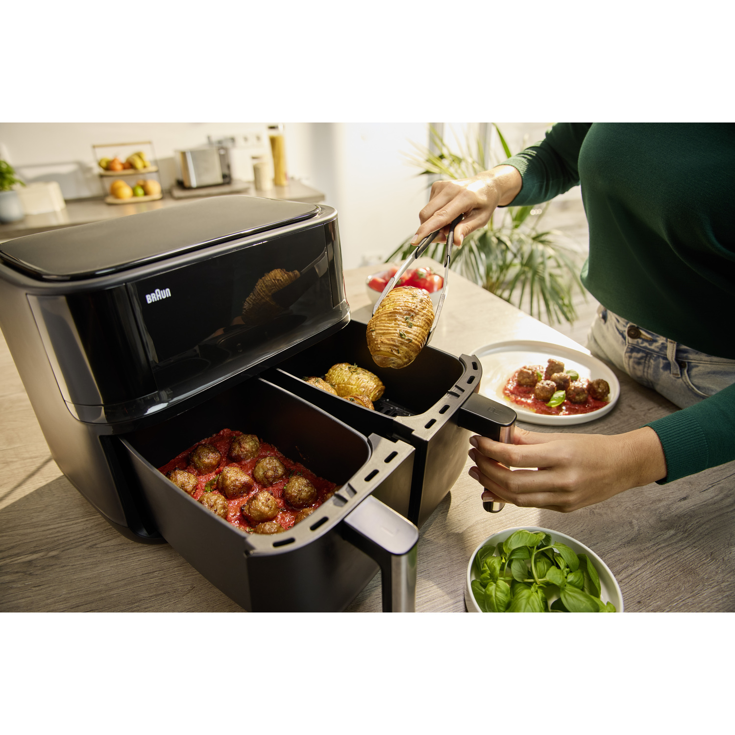 Een persoon gebruikt een tang om eten uit een zwarte airfryer te halen. Er zijn ook gehaktballen, basilicum en een bord met eten te zien.
