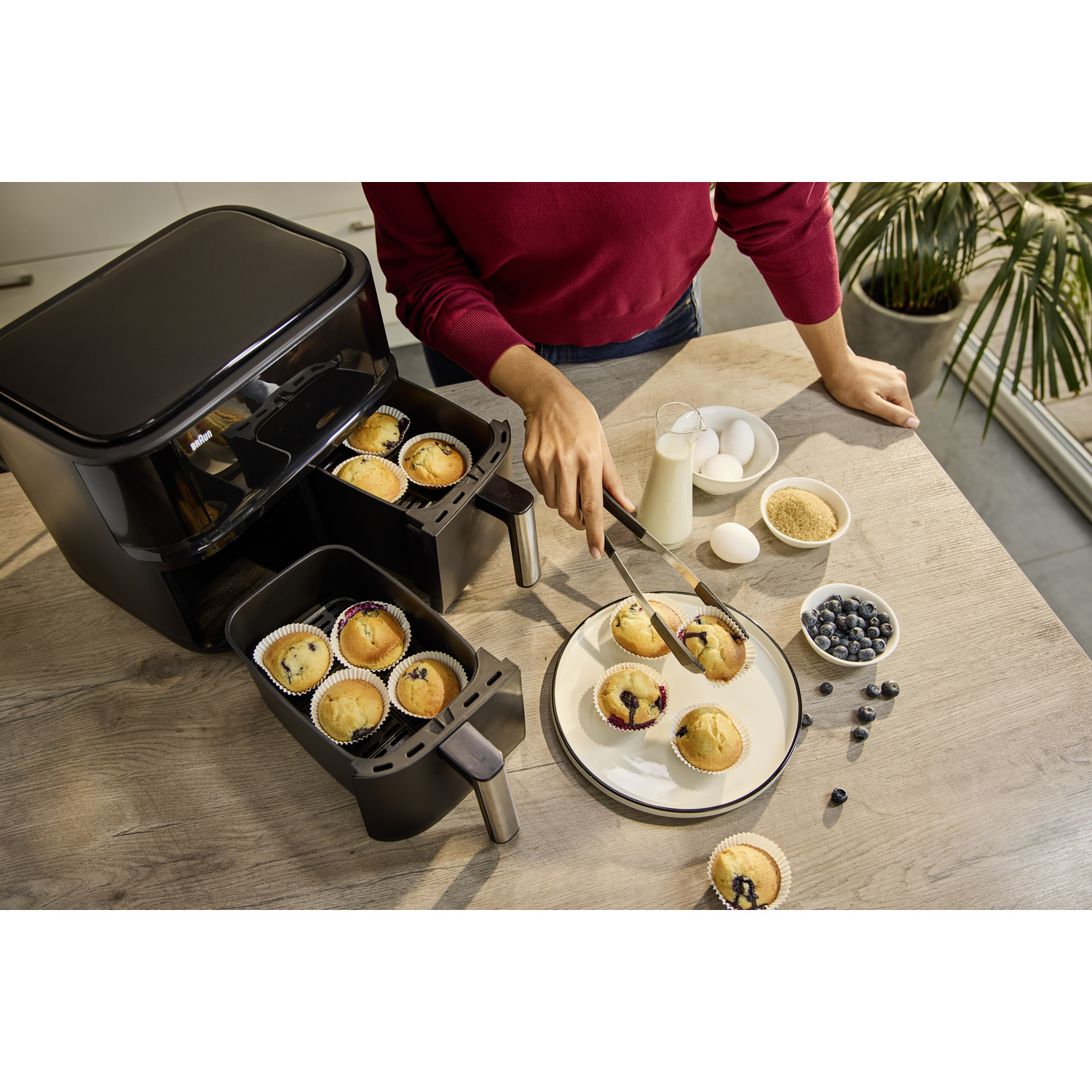 Persoon verwijdert muffins uit een airfryer. Ingrediënten als eieren, melk, blauwe bessen en suiker staan op tafel.