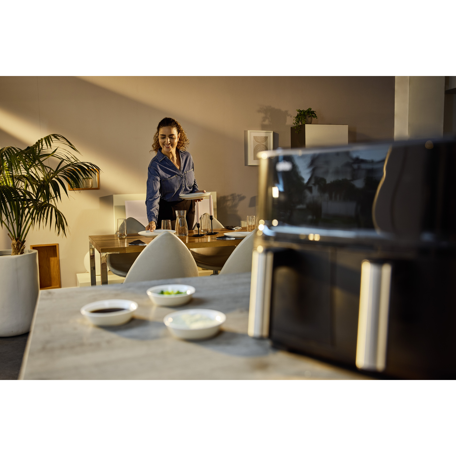 Een vrouw dekt de tafel. Een zwarte airfryer staat op de voorgrond. Kleine schaaltjes met sauzen staan op tafel.