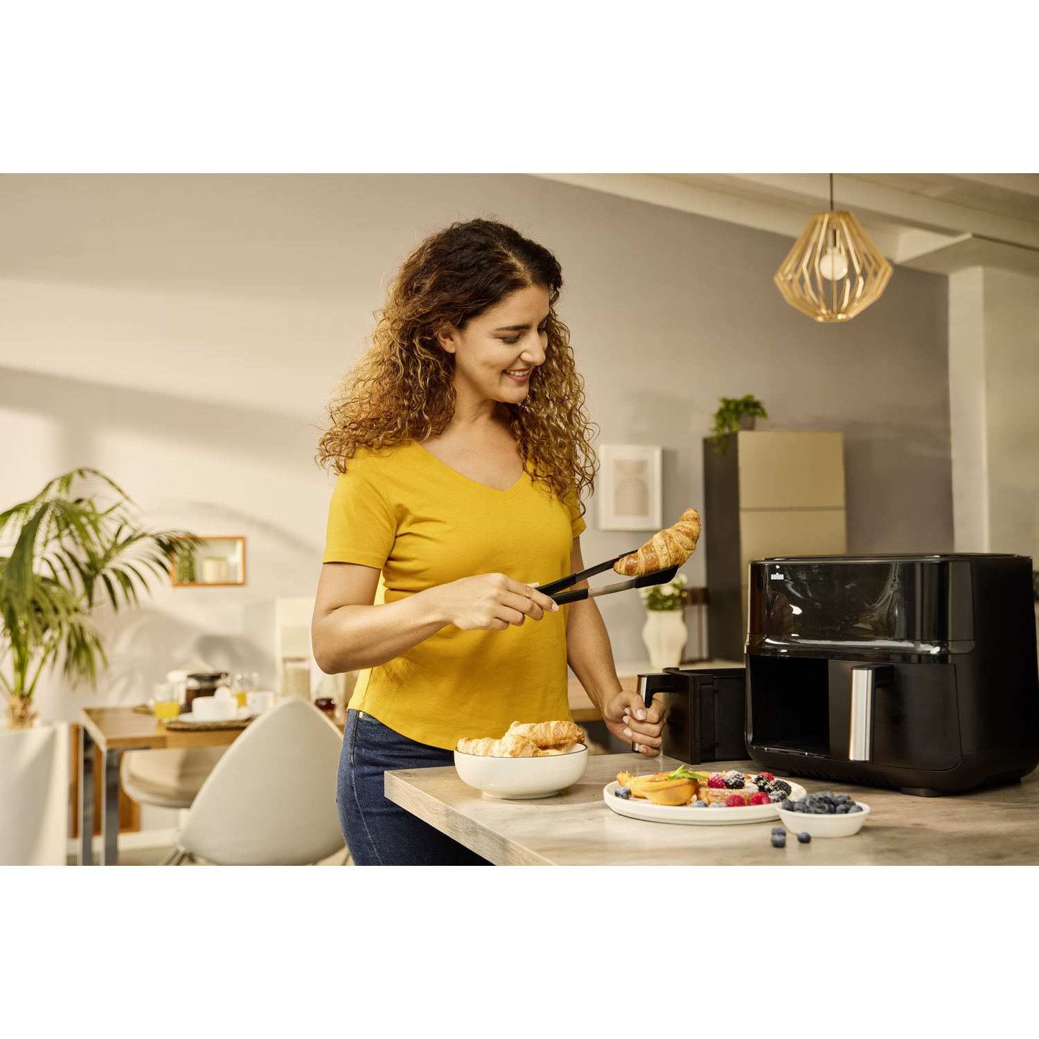 Vrouw met tang houdt croissant vast. Een zwarte airfryer en ontbijtbord op het aanrecht. Krullend haar, gele shirt.