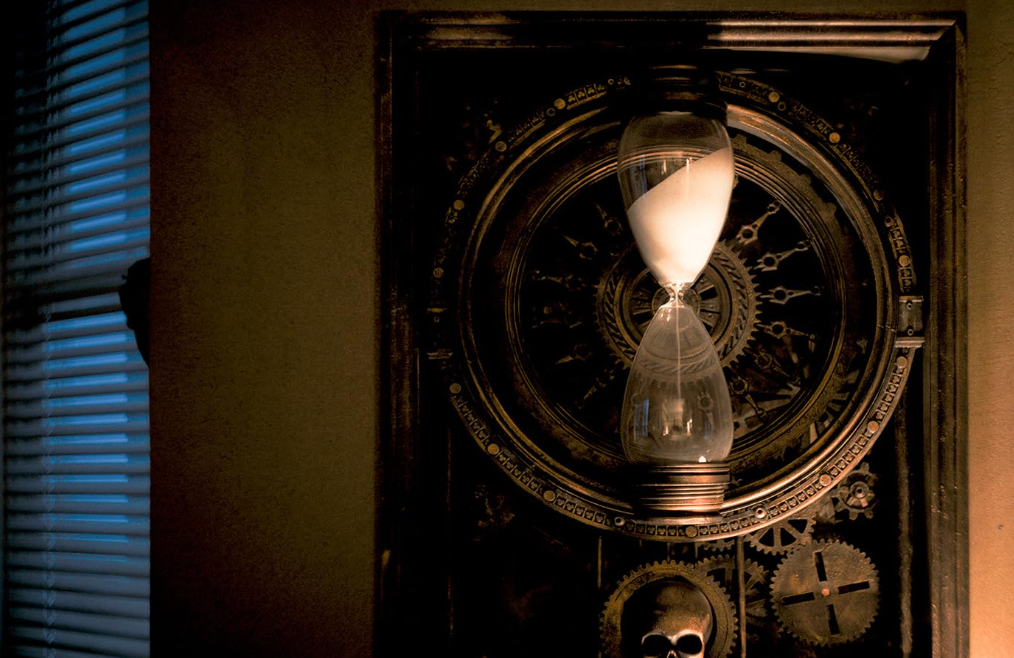 Eine Sanduhr an einer Wand mit Zahnrädern und einem Totenkopf, beleuchtet von Fensterlicht.