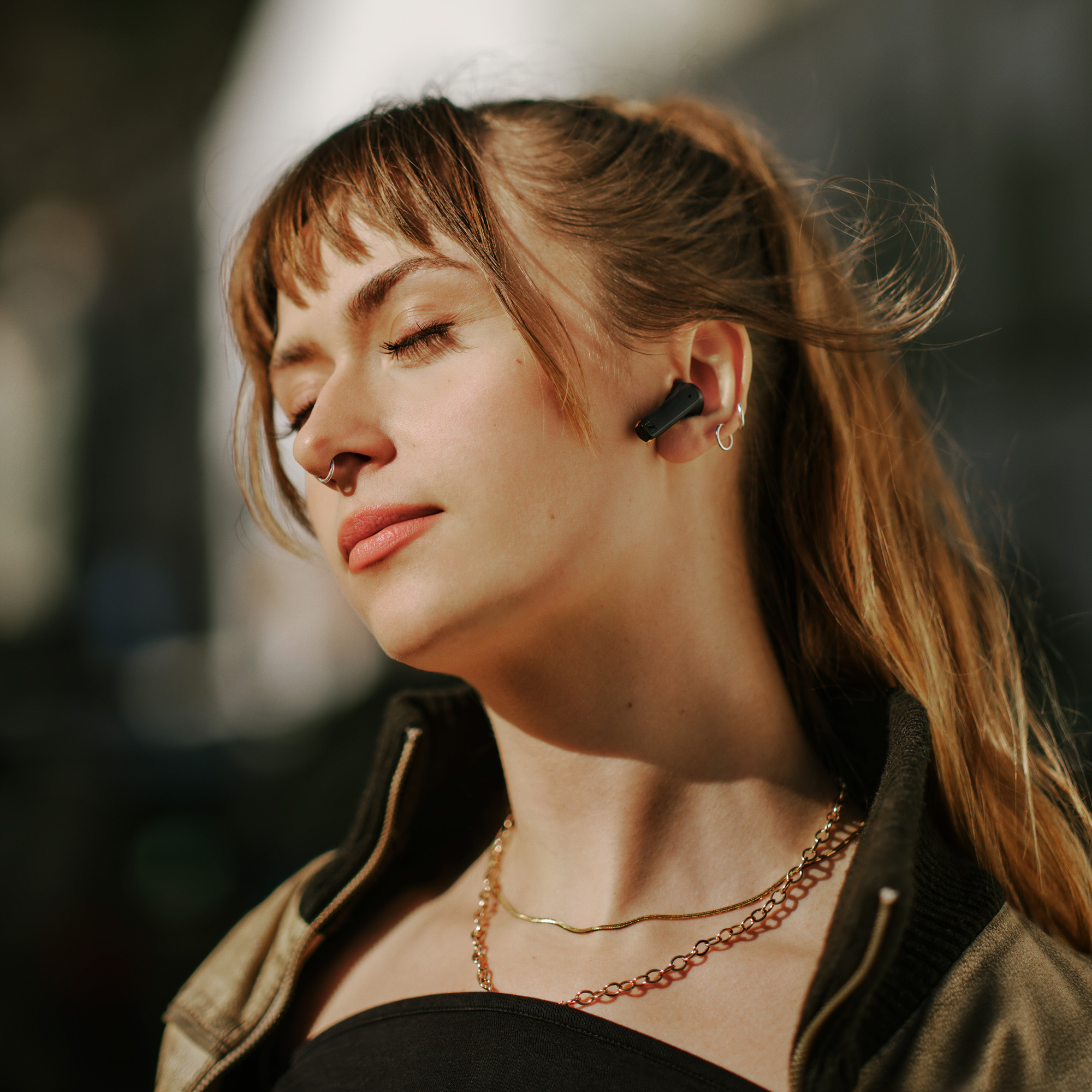 Femme avec écouteurs, yeux fermés, collier doré. Arrière-plan flou avec soleil.