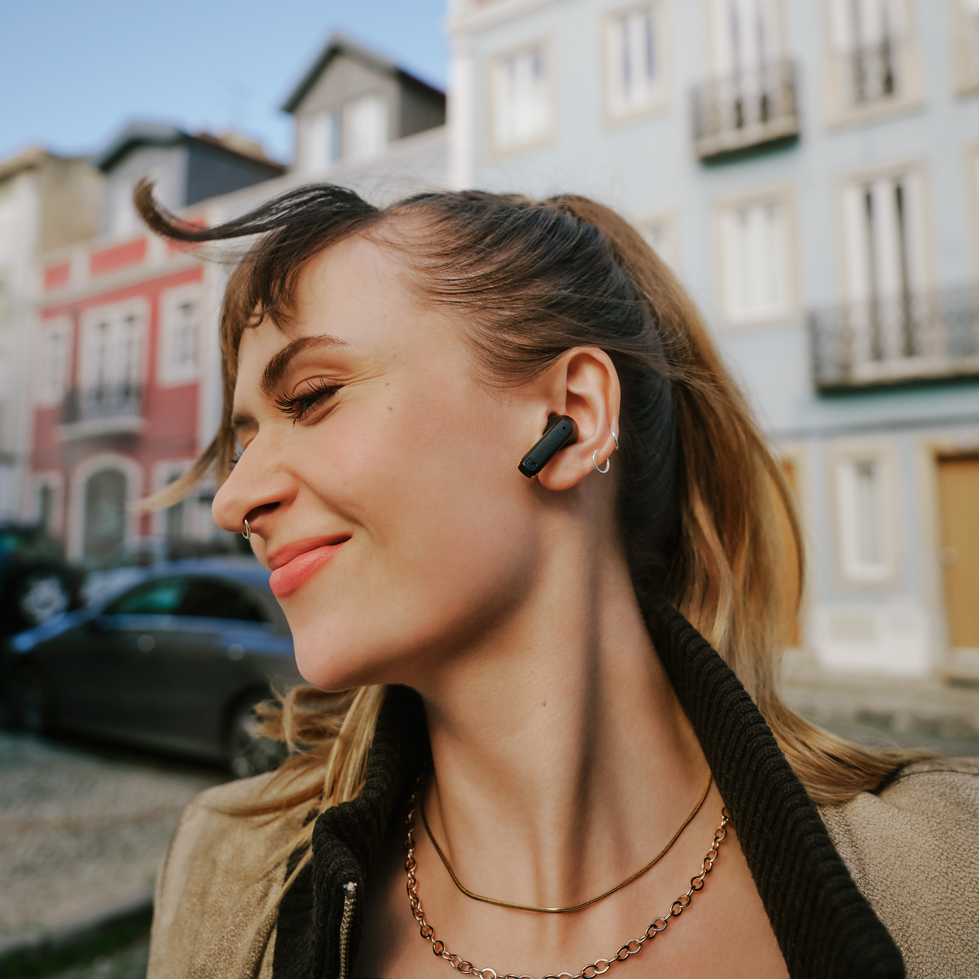 Femme souriante, écouteurs et collier doré, en milieu urbain avec des bâtiments.