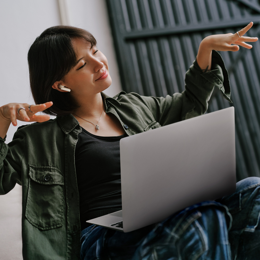 Femme avec des écouteurs, ordinateur portable, souriante, bras levés. Elle porte une veste et un jean.