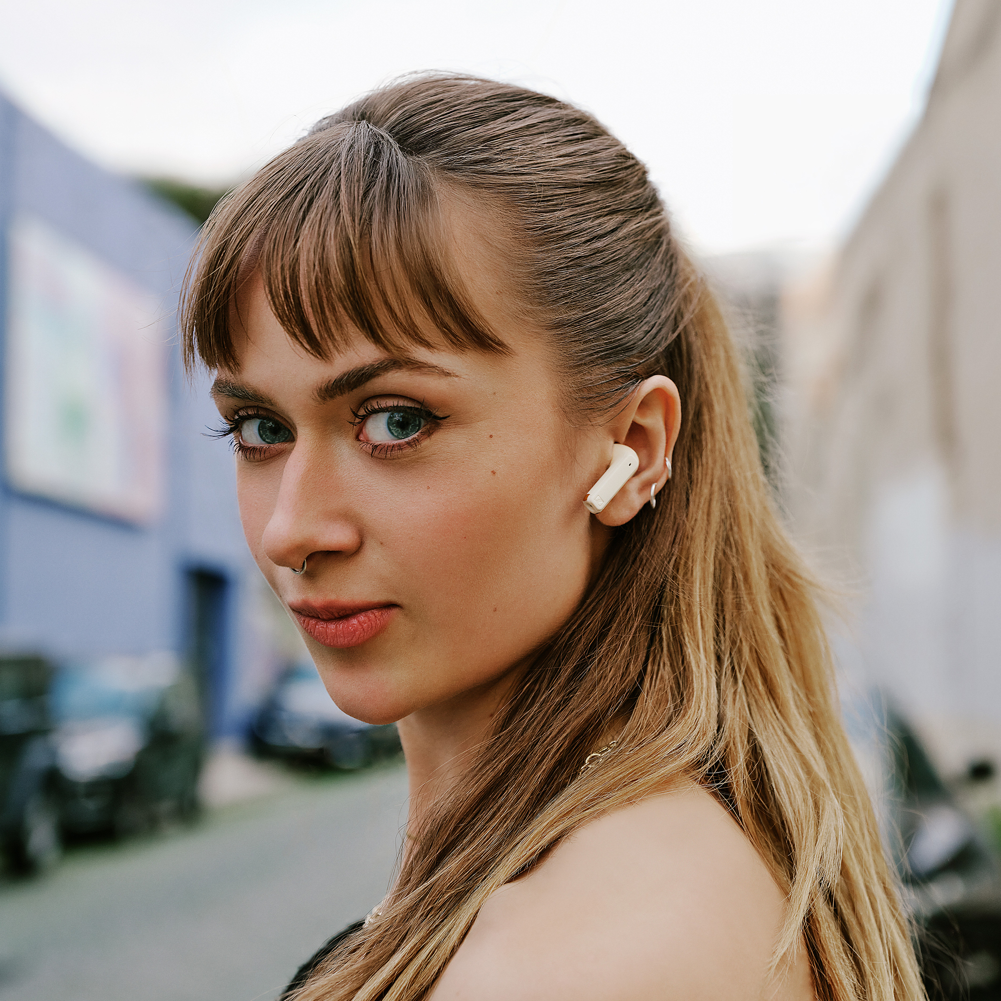 Femme avec des écouteurs, regarde la caméra, arrière-plan de rue. Elle porte un piercing au nez, cheveux blonds.