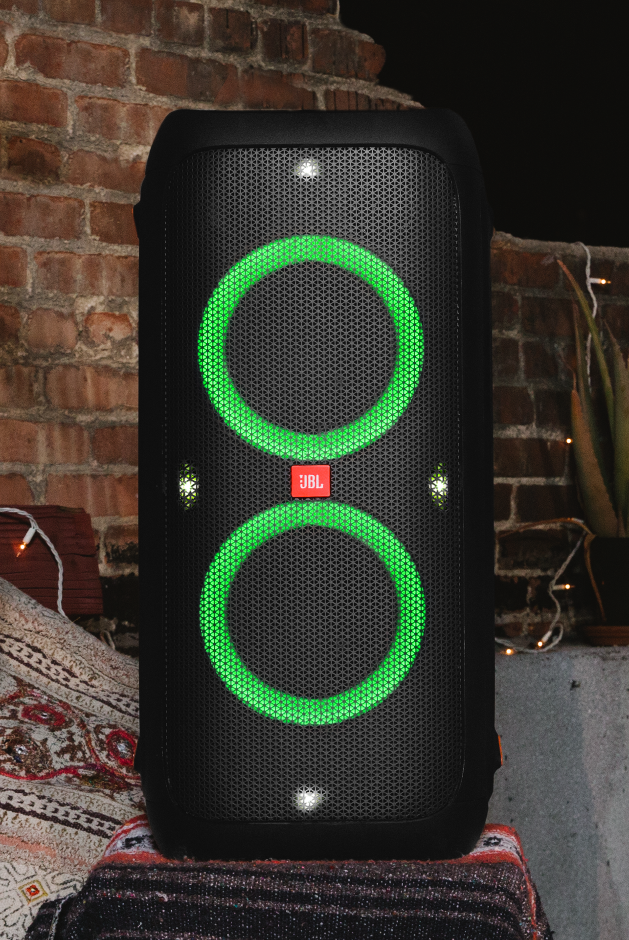 Schwarzer JBL-Lautsprecher mit grün leuchtenden Ringen. JBL-Logo in der Mitte. Ziegelwand im Hintergrund.