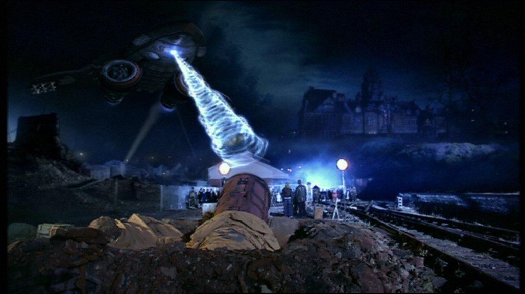UFO feuert Energie auf die Erde. Menschen schauen zu. Im Hintergrund ein Gebäude und Bahngleise. Nachtszene.