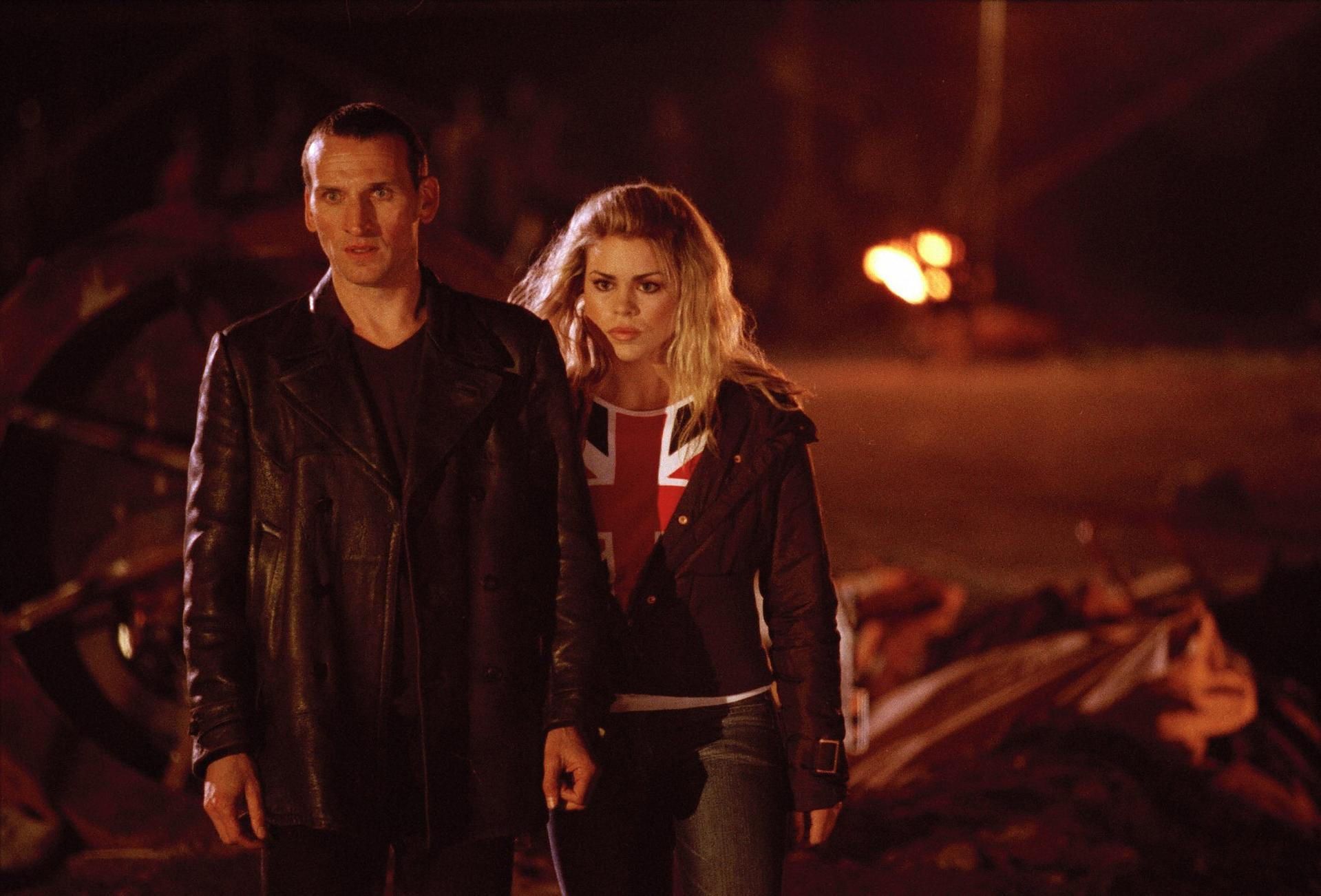Christopher Eccleston und Billie Piper stehen zusammen in einem schwach beleuchteten Bereich. Der Hintergrund ist verschwommen.