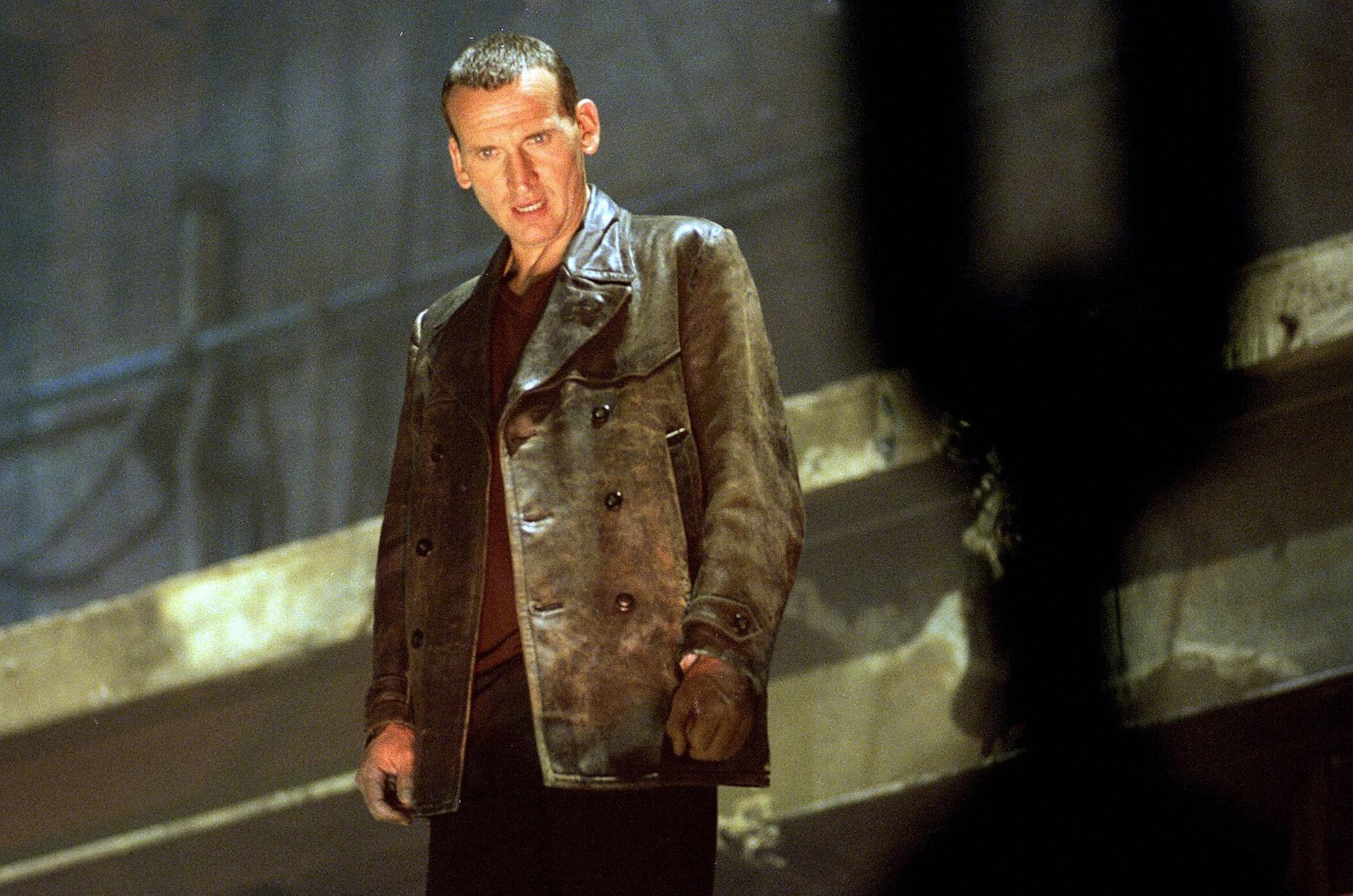 Christopher Eccleston in einem Ledermantel, der in einer dunklen, industriellen Umgebung steht.