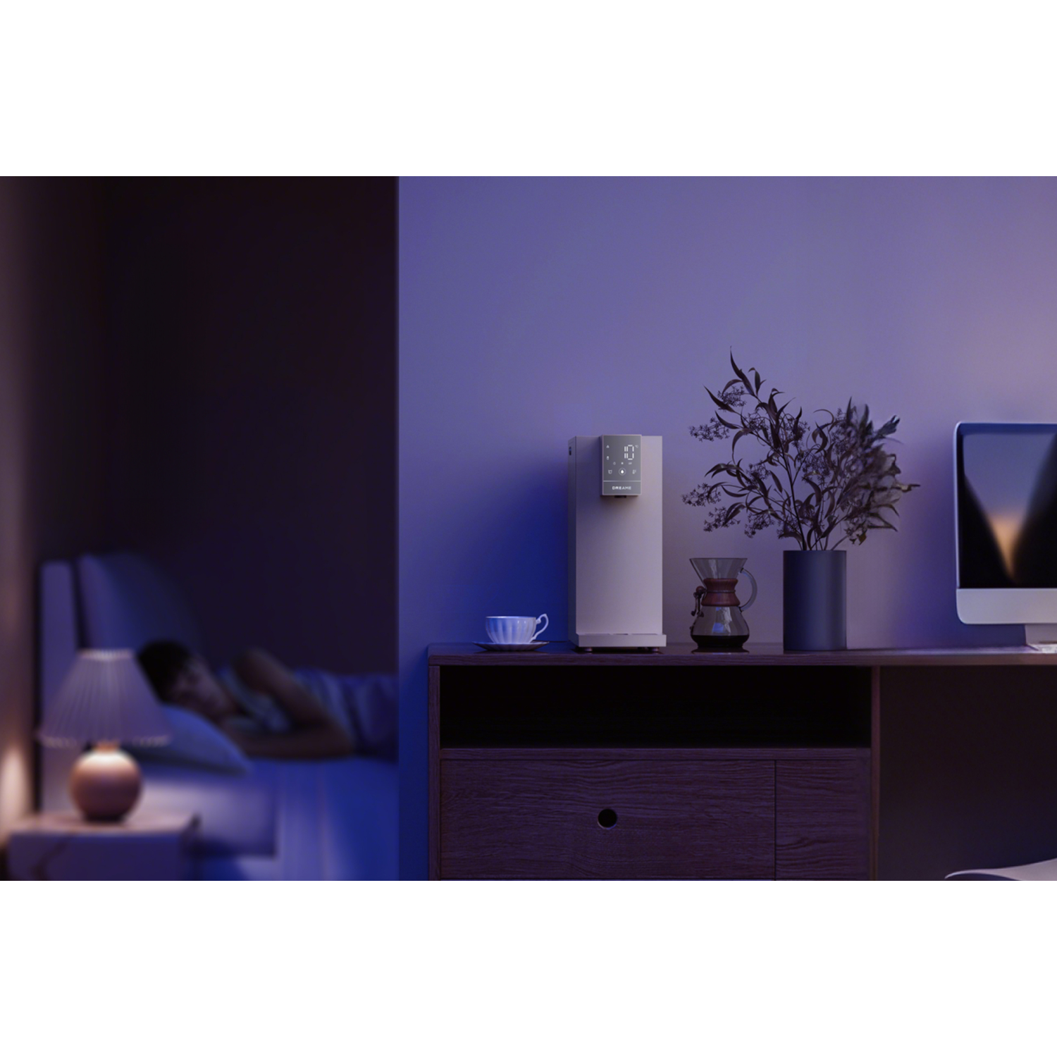 Een slaapkamer met een persoon in bed. Lamp, waterdispenser en koffiezetapparaat zijn zichtbaar.