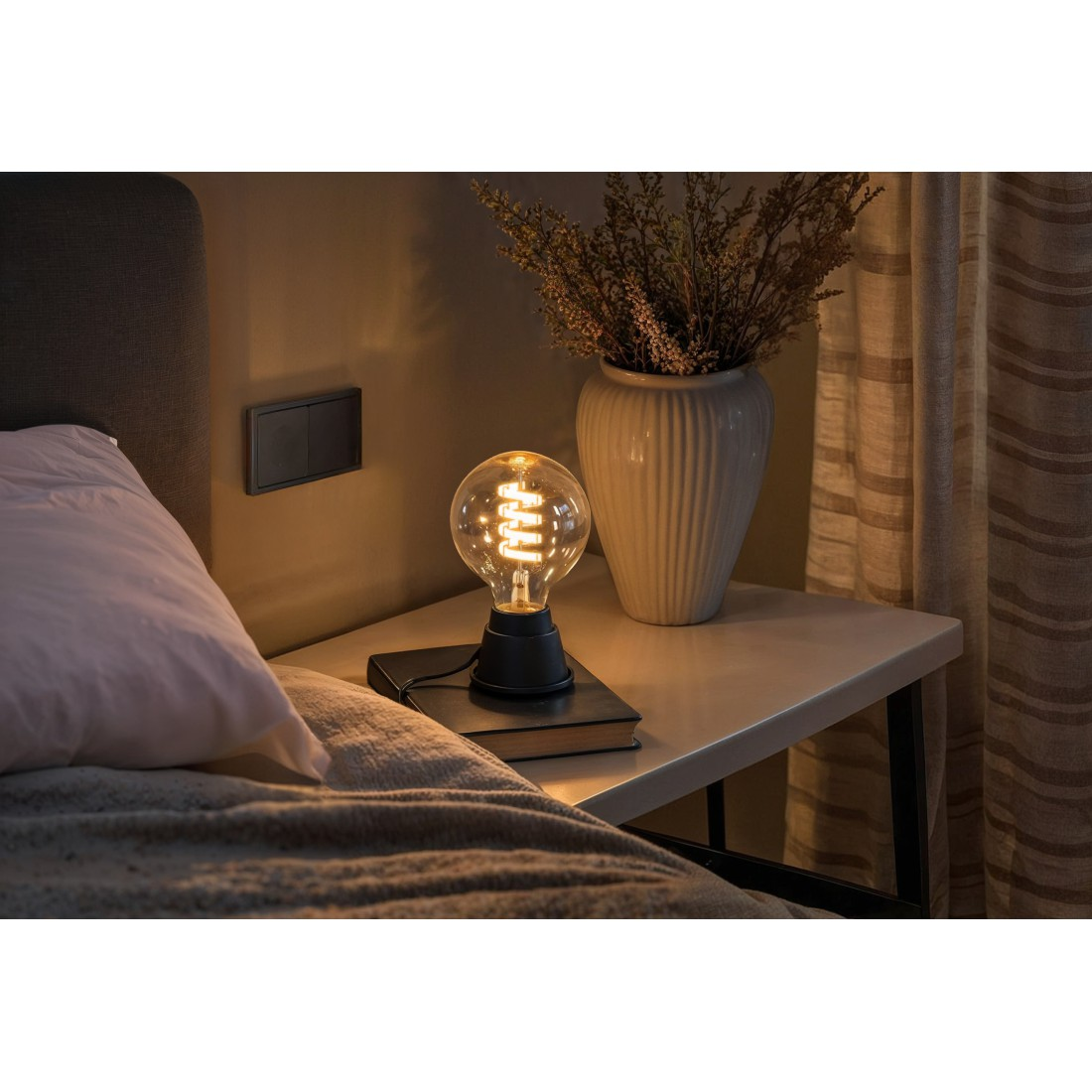 Nachttisch mit Lampe, Büchern und Vase mit Blumen. Der Raum ist dunkel.