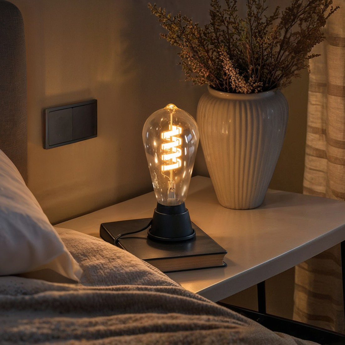 Eine beleuchtete Glühbirne auf einem Nachttisch neben einem Bett, mit einem Buch und einer Vase.
