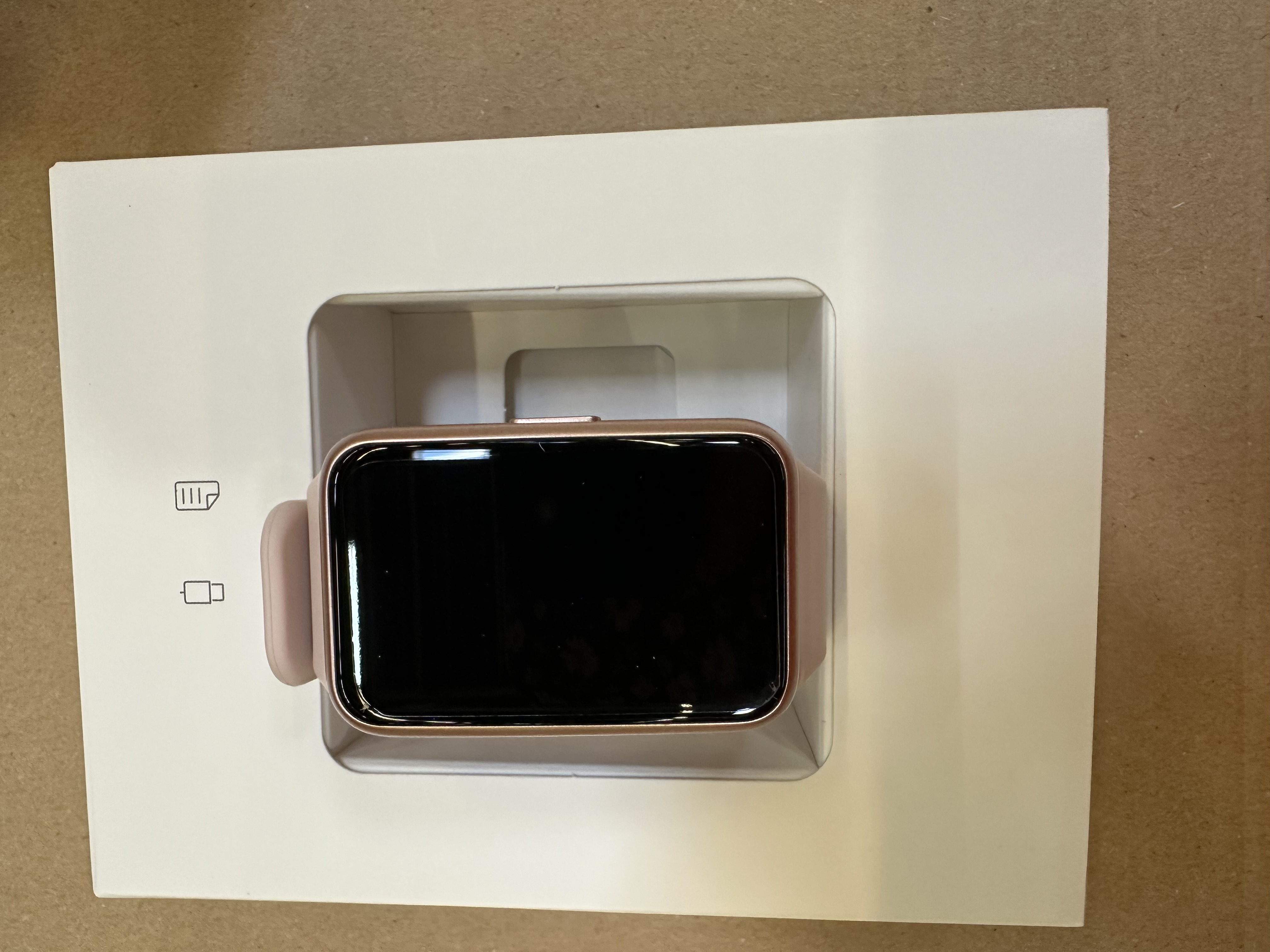 Różowy smartwatch w białym pudełku. Ekran jest czarny. Obok zegarka znajduje się karta.