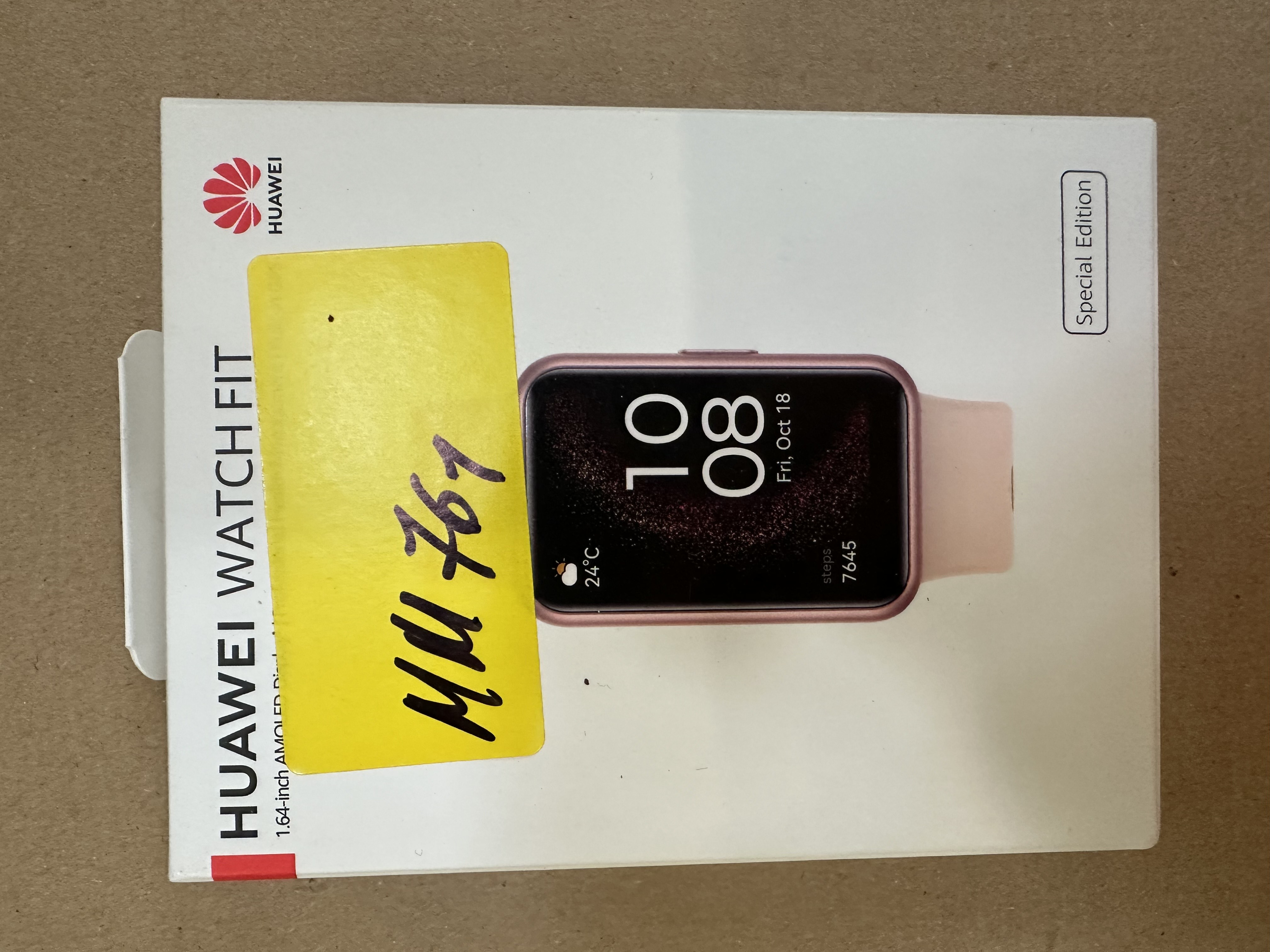 Smartwatch w pudełku. Ekran wyświetla datę i godzinę. Żółta naklejka znajduje się na pudełku.