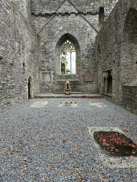 Intérieur d'une vieille église en pierre. Une fenêtre en arc, une chaise dorée et une porte sont présentes. Le sol est recouvert de gravier.