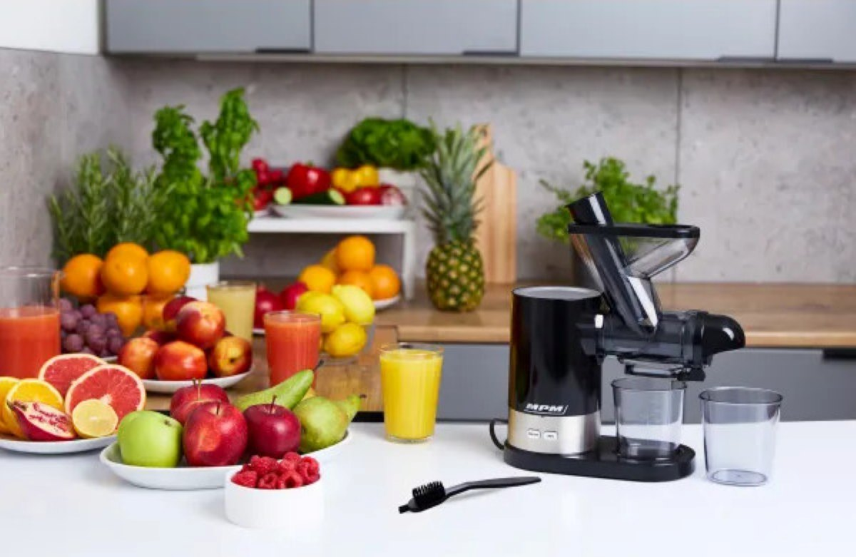 Scena kuchenna: Wyciskarka i owoce, szklanki soku na białej powierzchni.