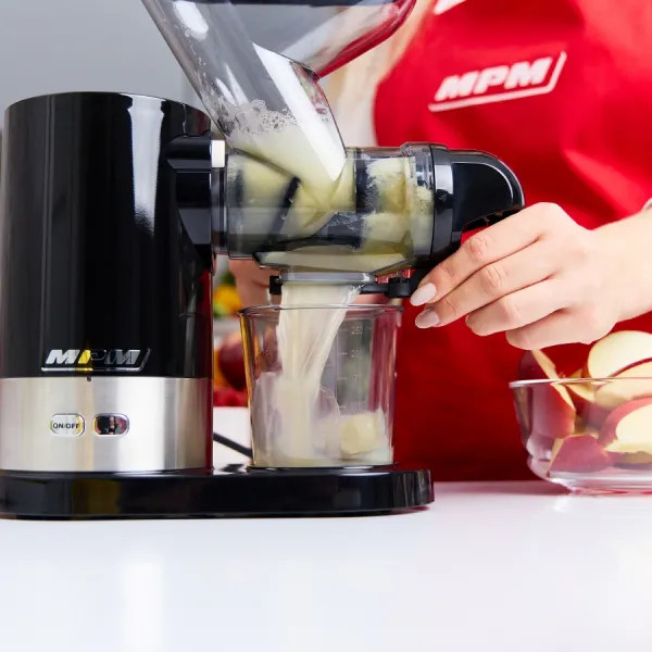 Sok wlewa się z wyciskarki do szklanki. Pokrojone jabłka w misce. Osoba ma na sobie czerwony fartuch.