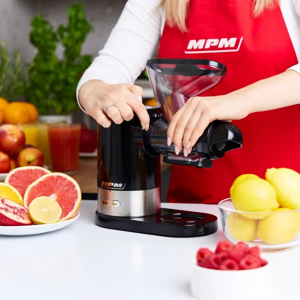 Osoba używa wyciskarki do soku. Plastry grejpfruta, cytryny i maliny na pierwszym planie. Widoczny czerwony fartuch.