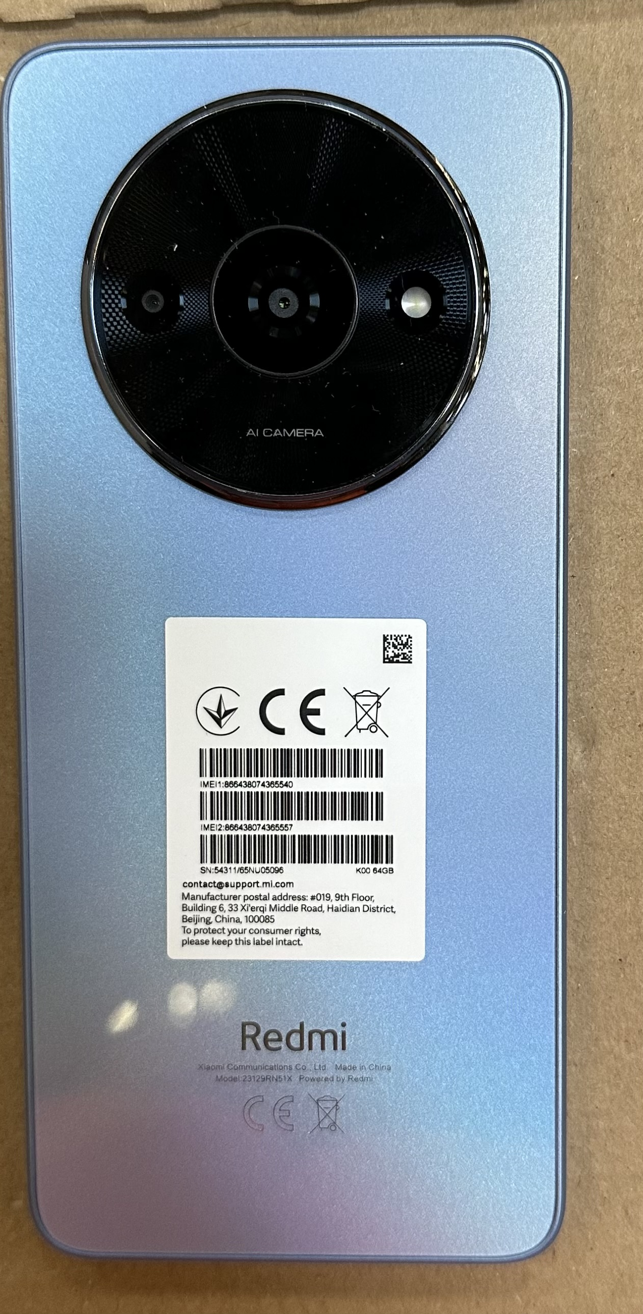 Tył niebieskiego telefonu Redmi z dużym czarnym modułem kamery. Biała etykieta z tekstem i kodami kreskowymi.