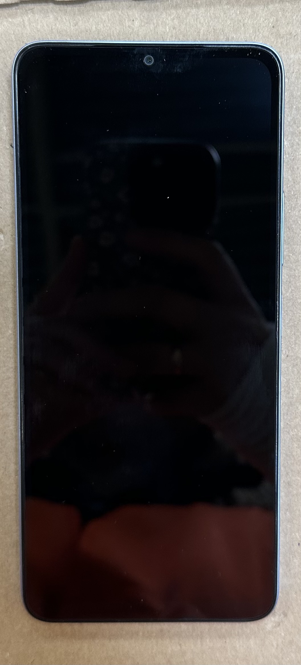 Czarny smartfon z ciemnym ekranem, przednim aparatem i odbiciem. Na jasnobrązowej powierzchni.