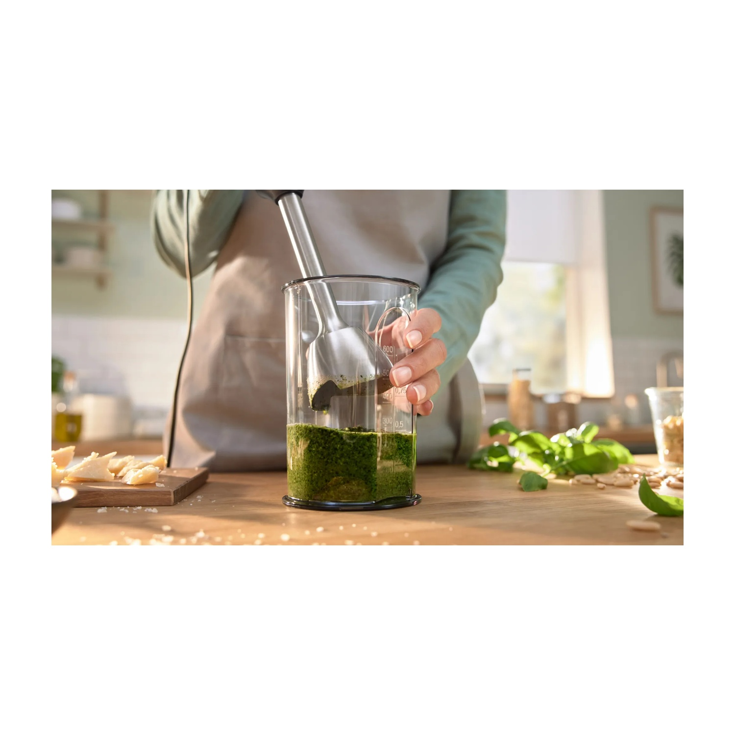 Un frullatore sta mescolando il pesto in un bicchiere. Ingredienti come formaggio e basilico sul tavolo.