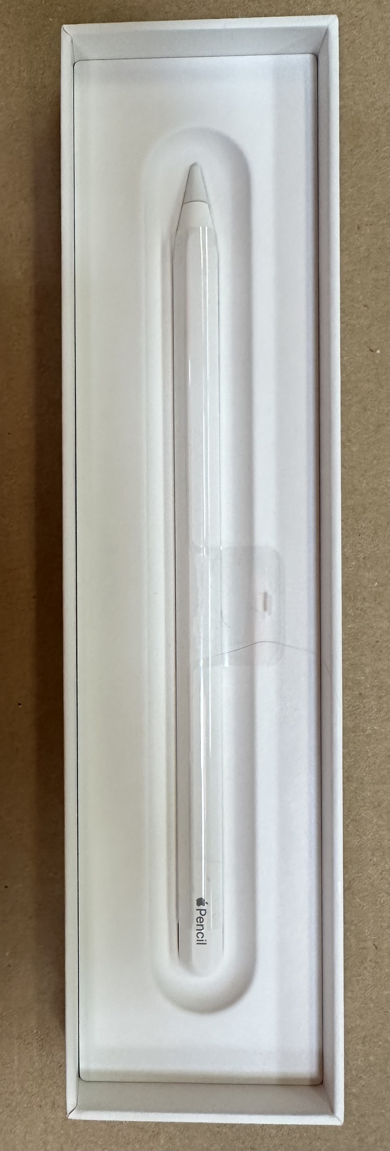 Białe pudełko z ołówkiem Apple w środku. Pudełko jest otwarte. Tłem jest brązowa powierzchnia.